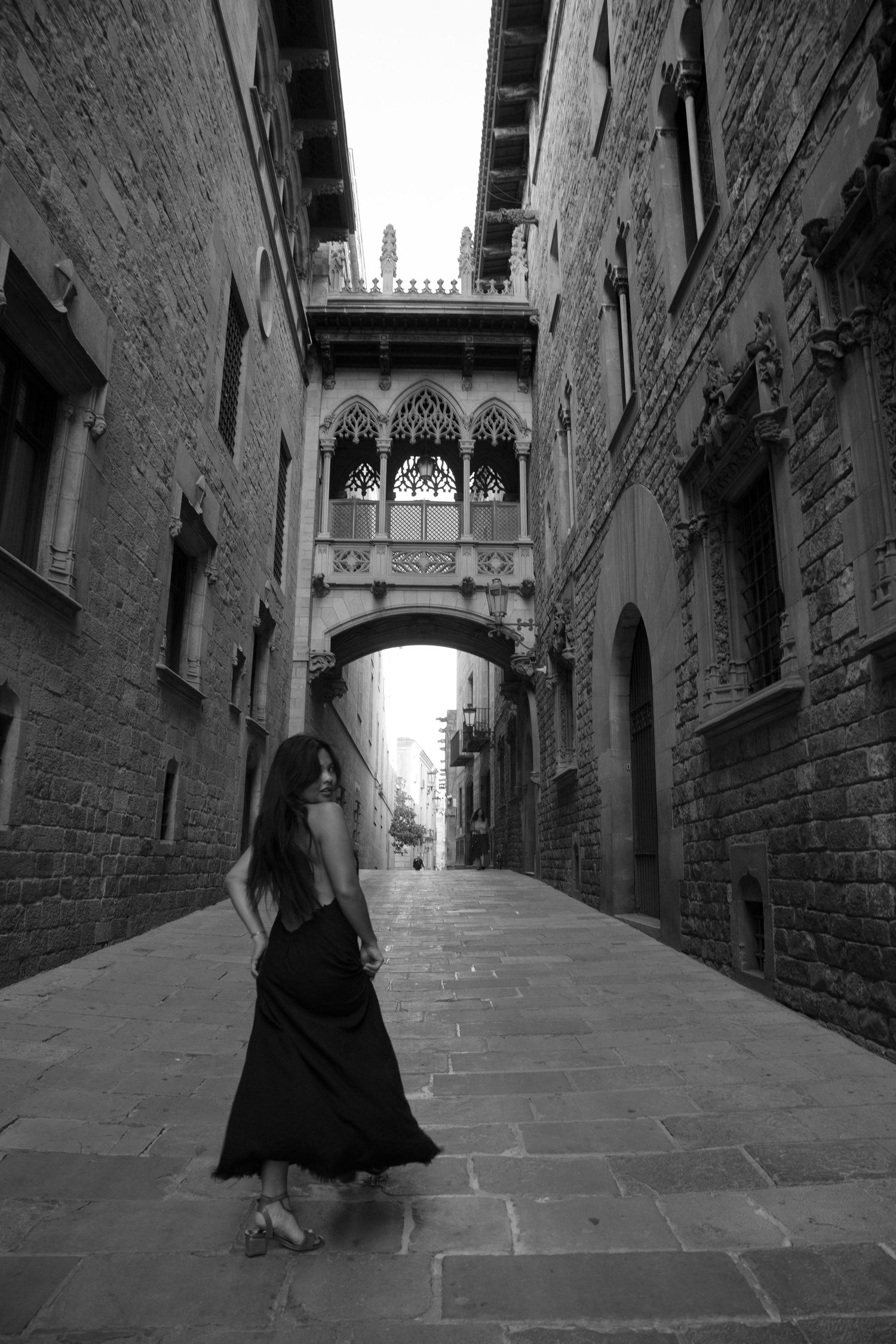 Woman in black dress walks cobblestone alley beneath Gothic arch bridge in historic Barcelona neighborhood.