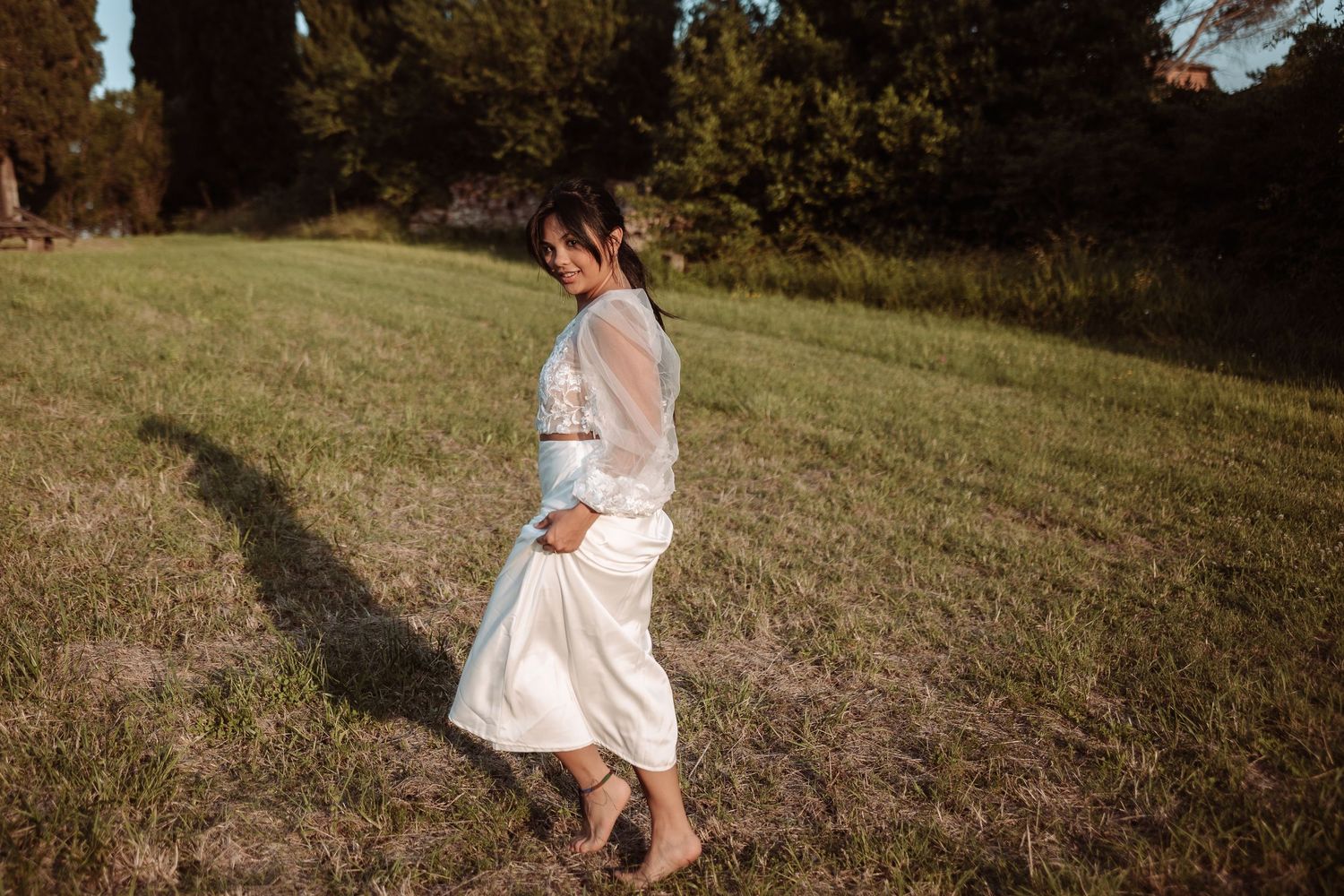 Figure in a flowing white dress walking through a sunlit grassy meadow.