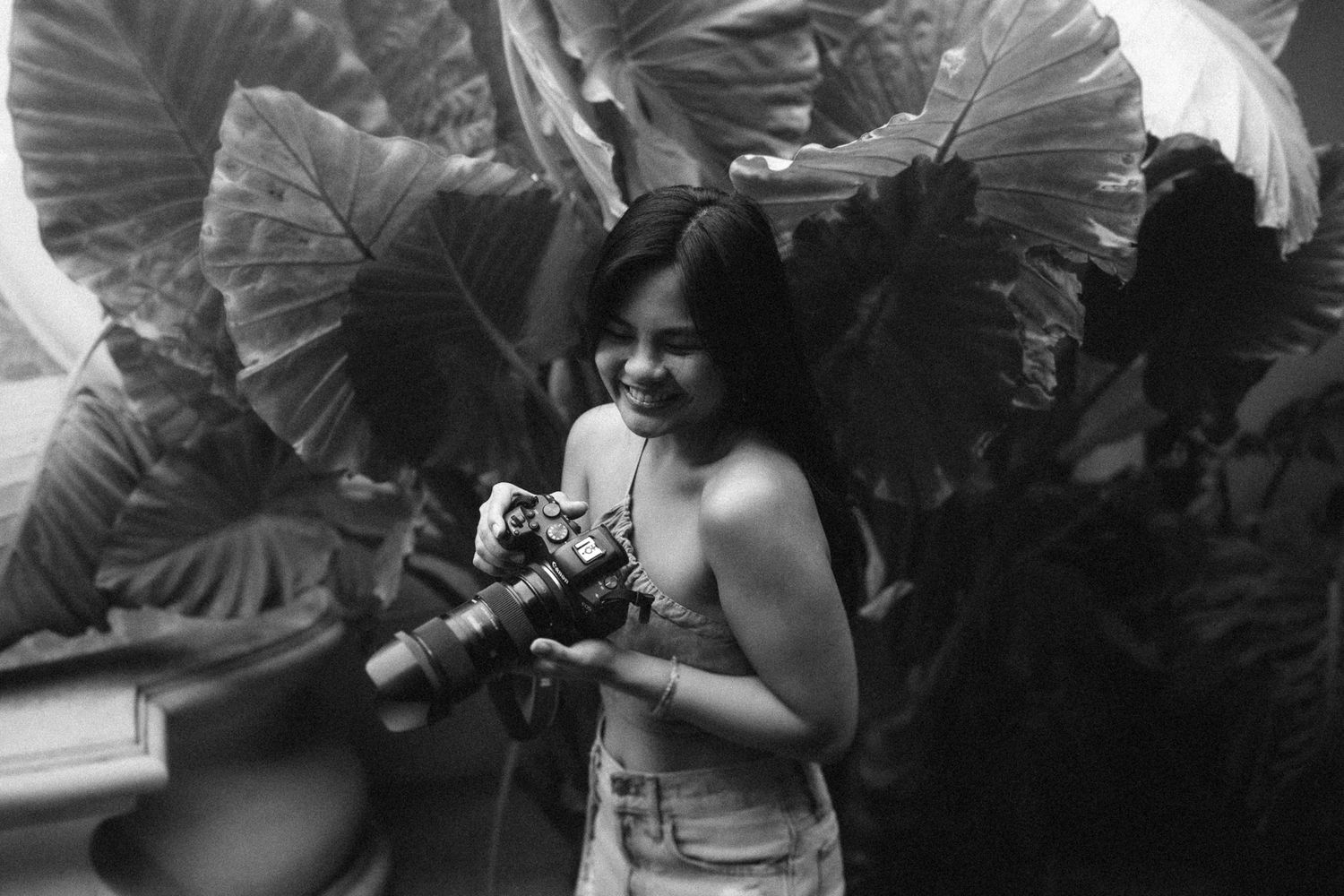 A photographer holds a vintage camera while standing among large tropical leaves in a black and white photograph.