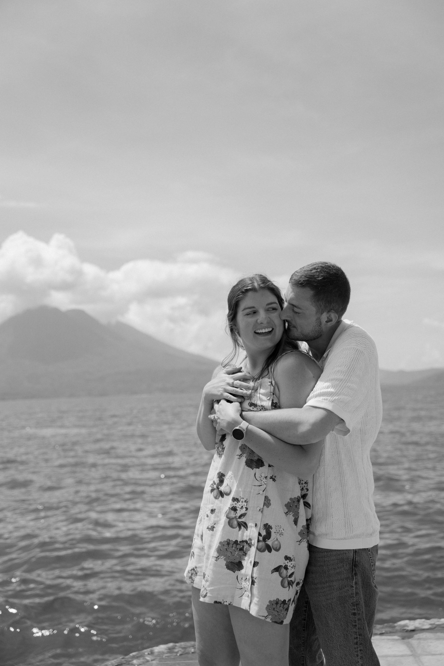 A couple shares a romantic moment by the ocean with mountains visible in the distance, captured in black and white.