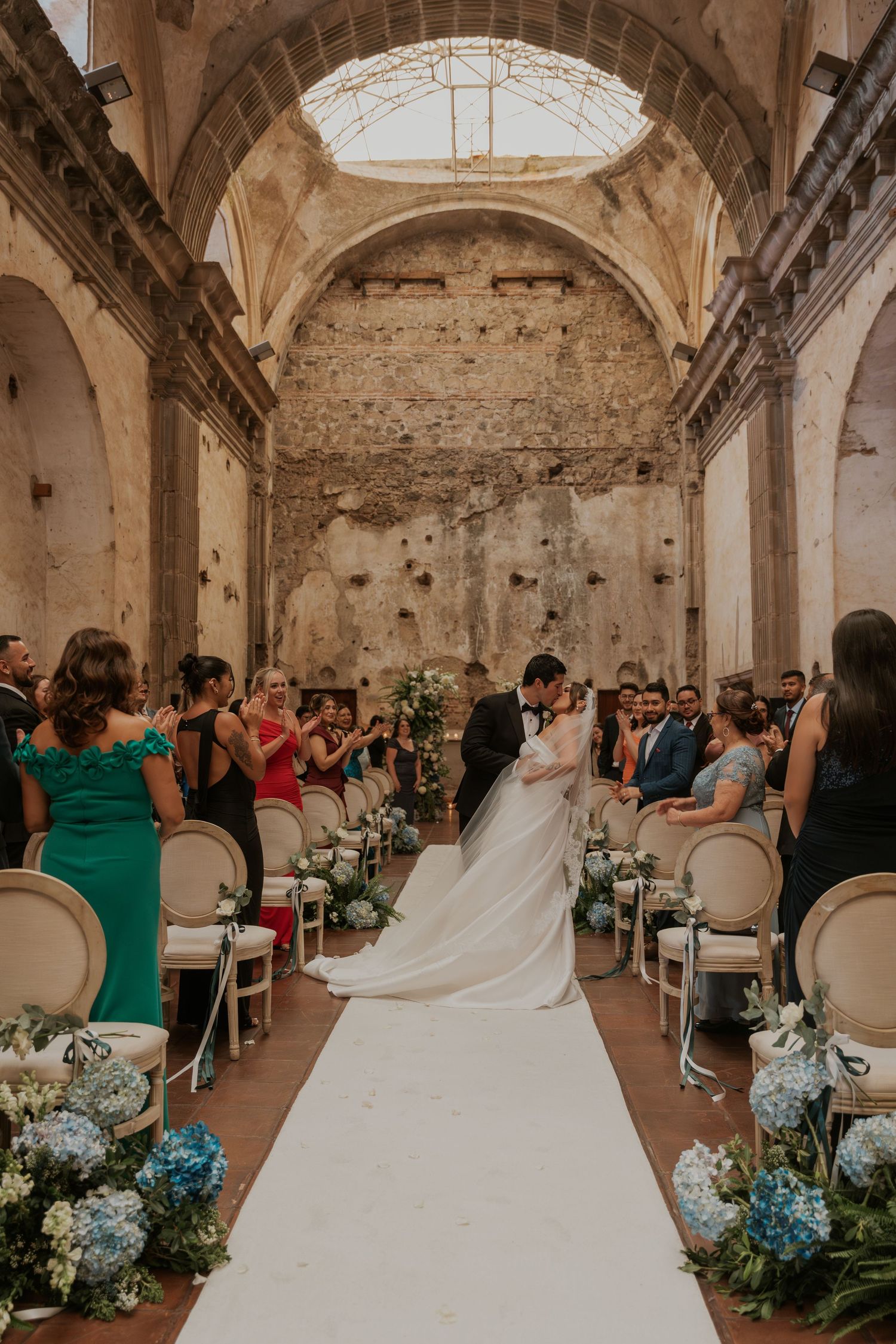 A wedding ceremony in a rustic stone chapel with white chairs and blue floral decorations along the aisle.
