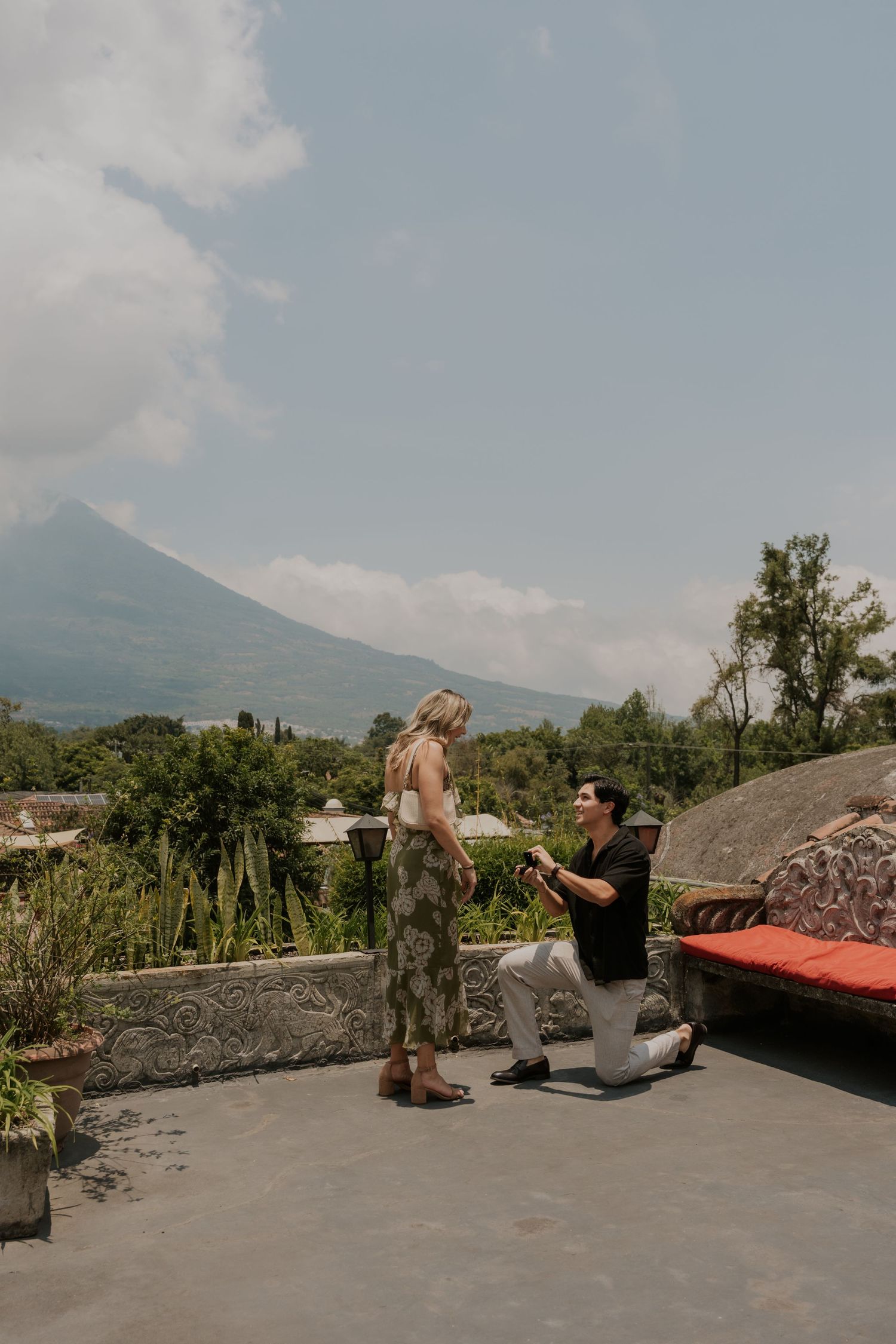 Man proposes on one knee to woman on terrace with lush greenery and volcano backdrop.