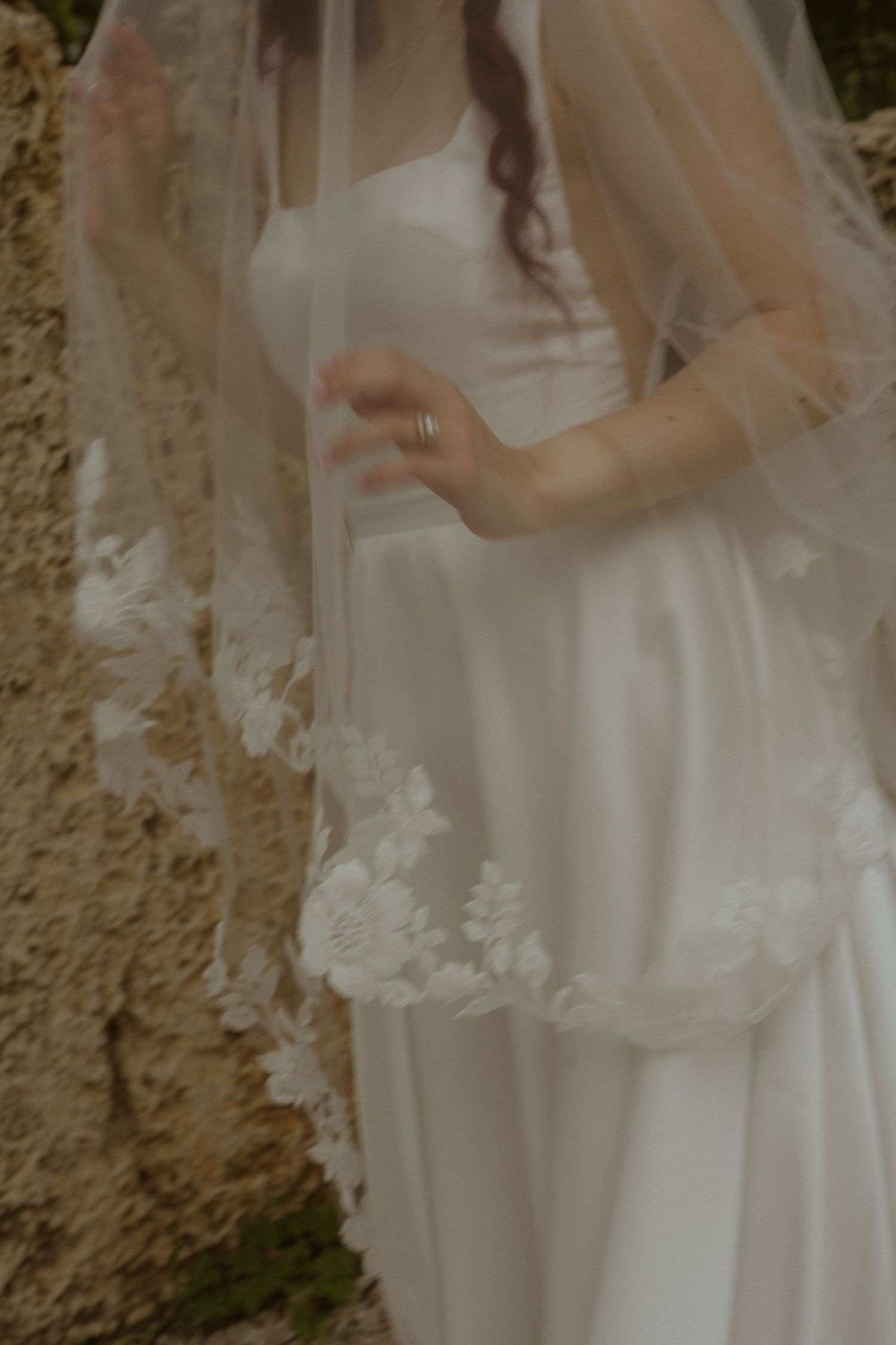 Bride in white satin gown with floral lace-trimmed veil outdoors near stone wall.
