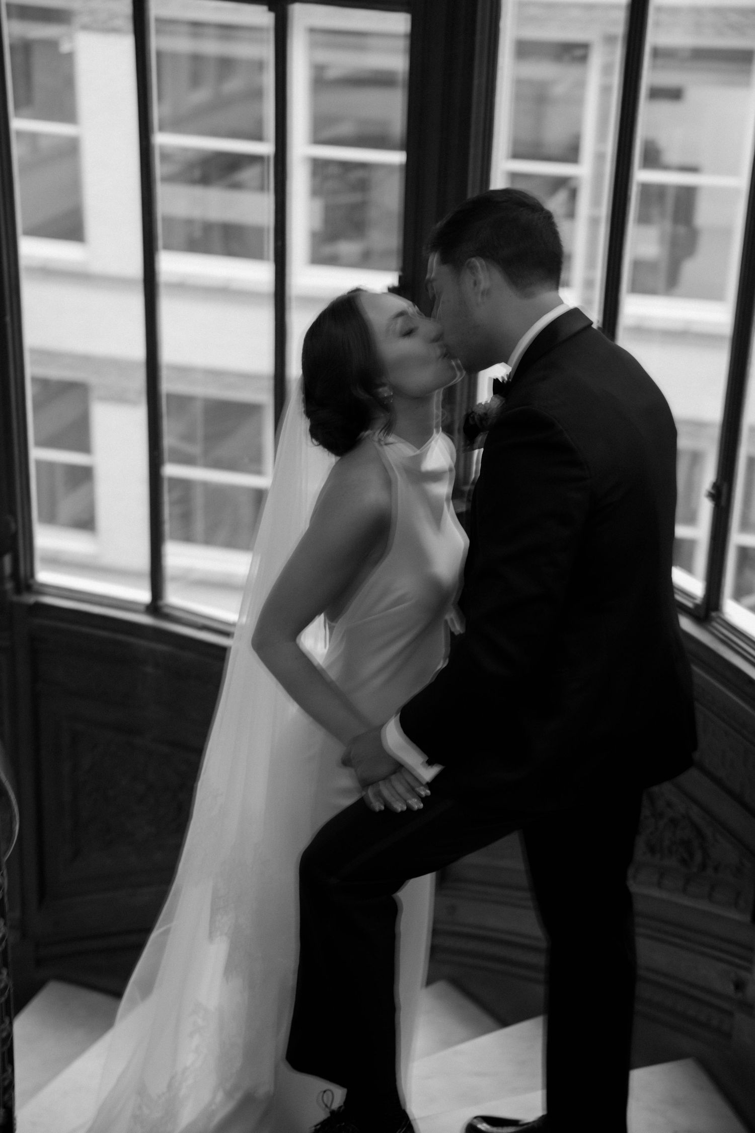 Romantic black and white wedding photo of a couple sharing a kiss by large windows.