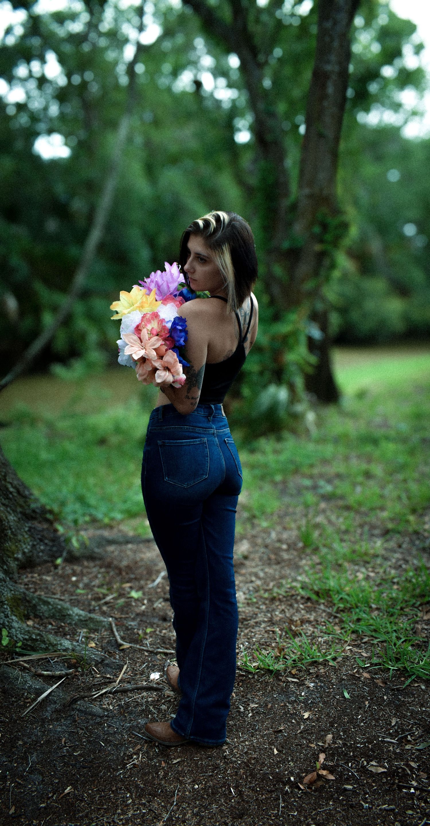 Woman in jeans and black top holds colorful flower bouquet in a lush green forest setting.