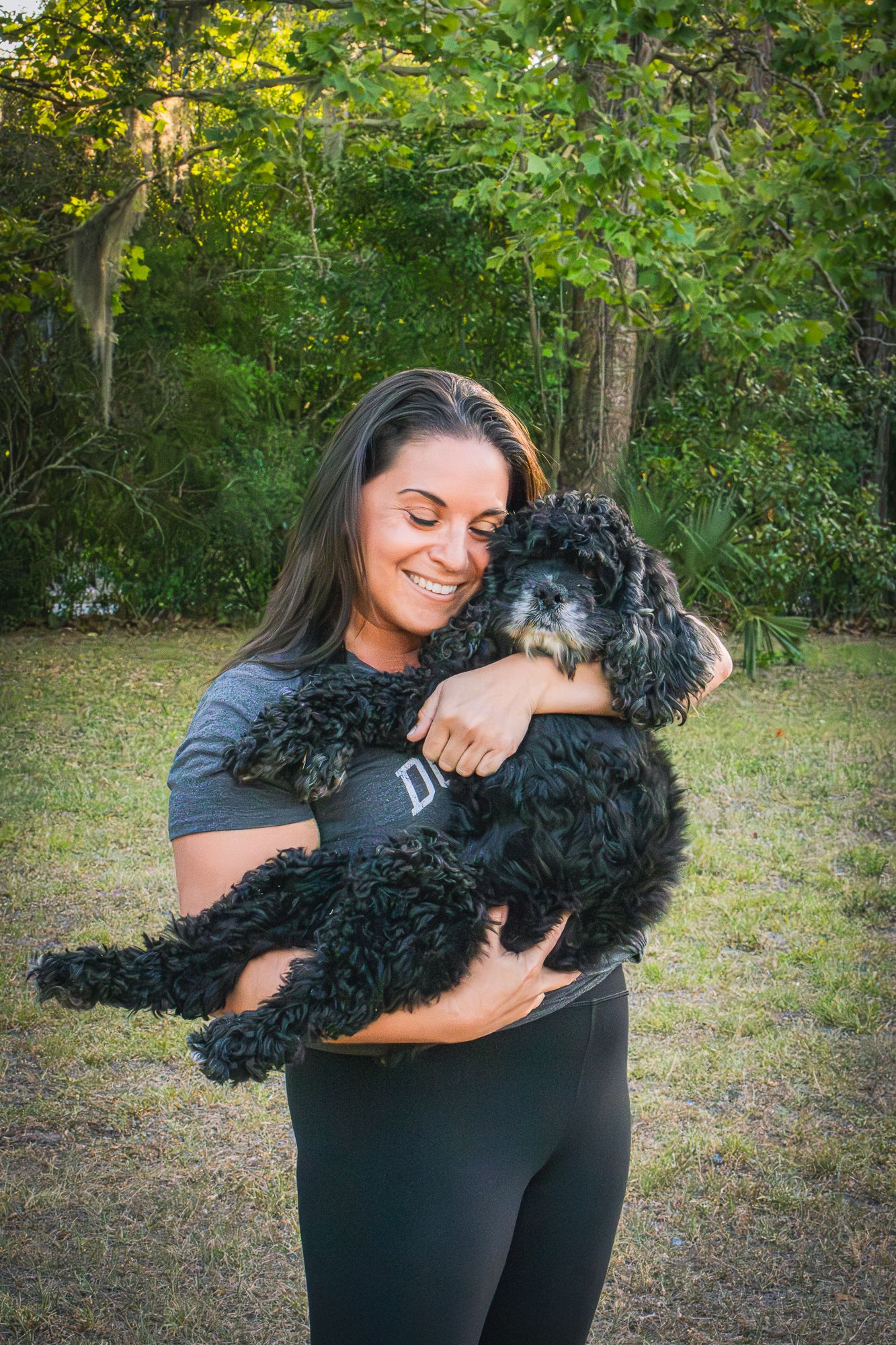 Smiling woman with dark hair hugging a fluffy black poodle dog outdoors in a green park.