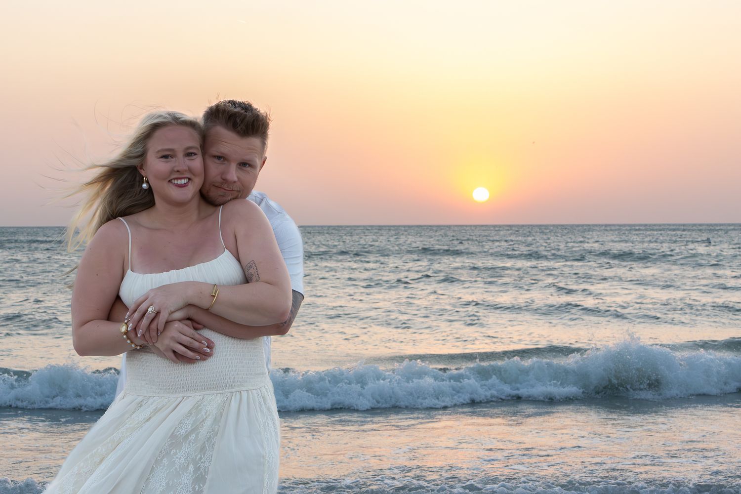 Happy couple embracing on a beach at sunset with golden pink sky reflecting over ocean waves.