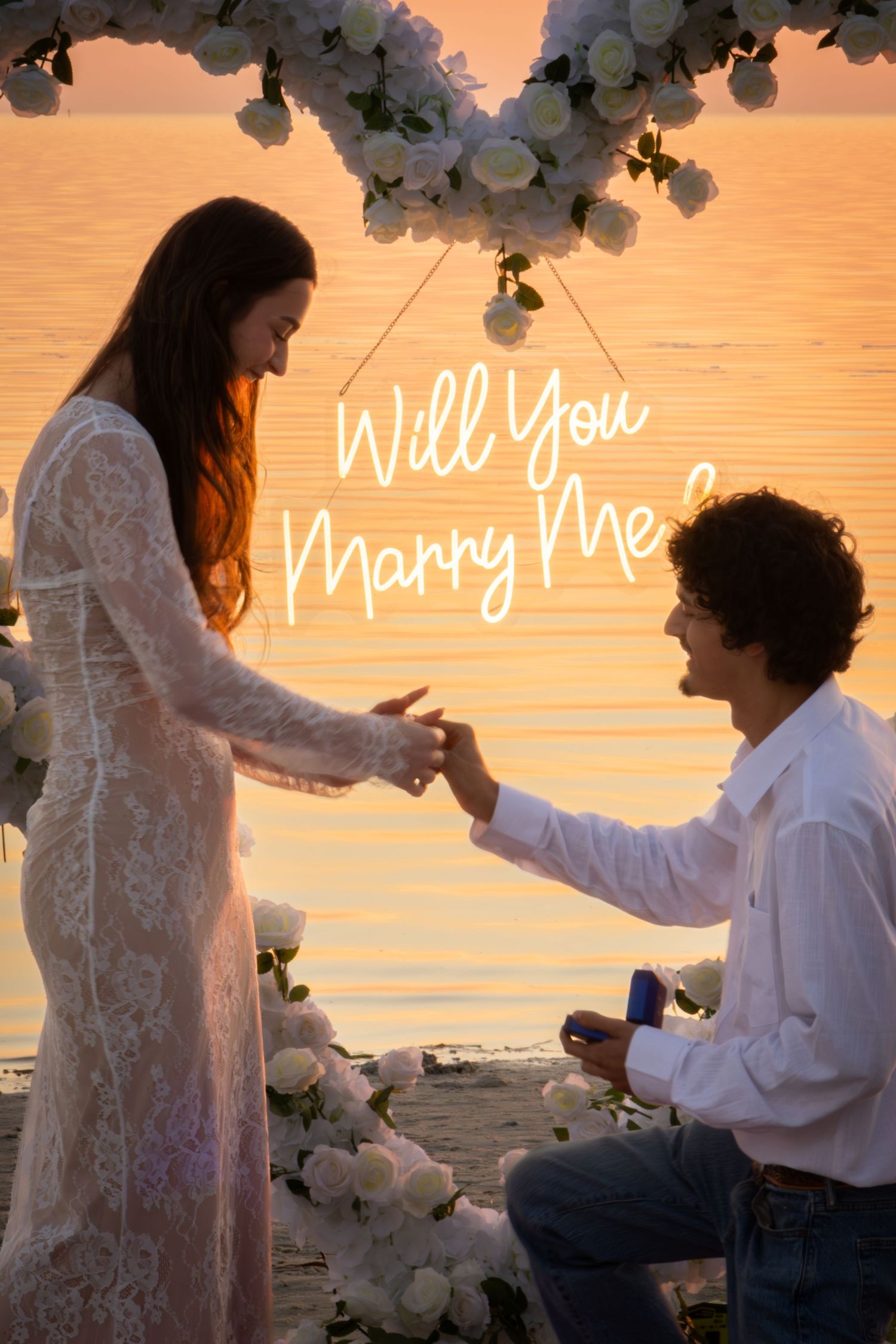 Romantic beach sunset marriage proposal with Will You Marry Me neon sign and floral arch.
