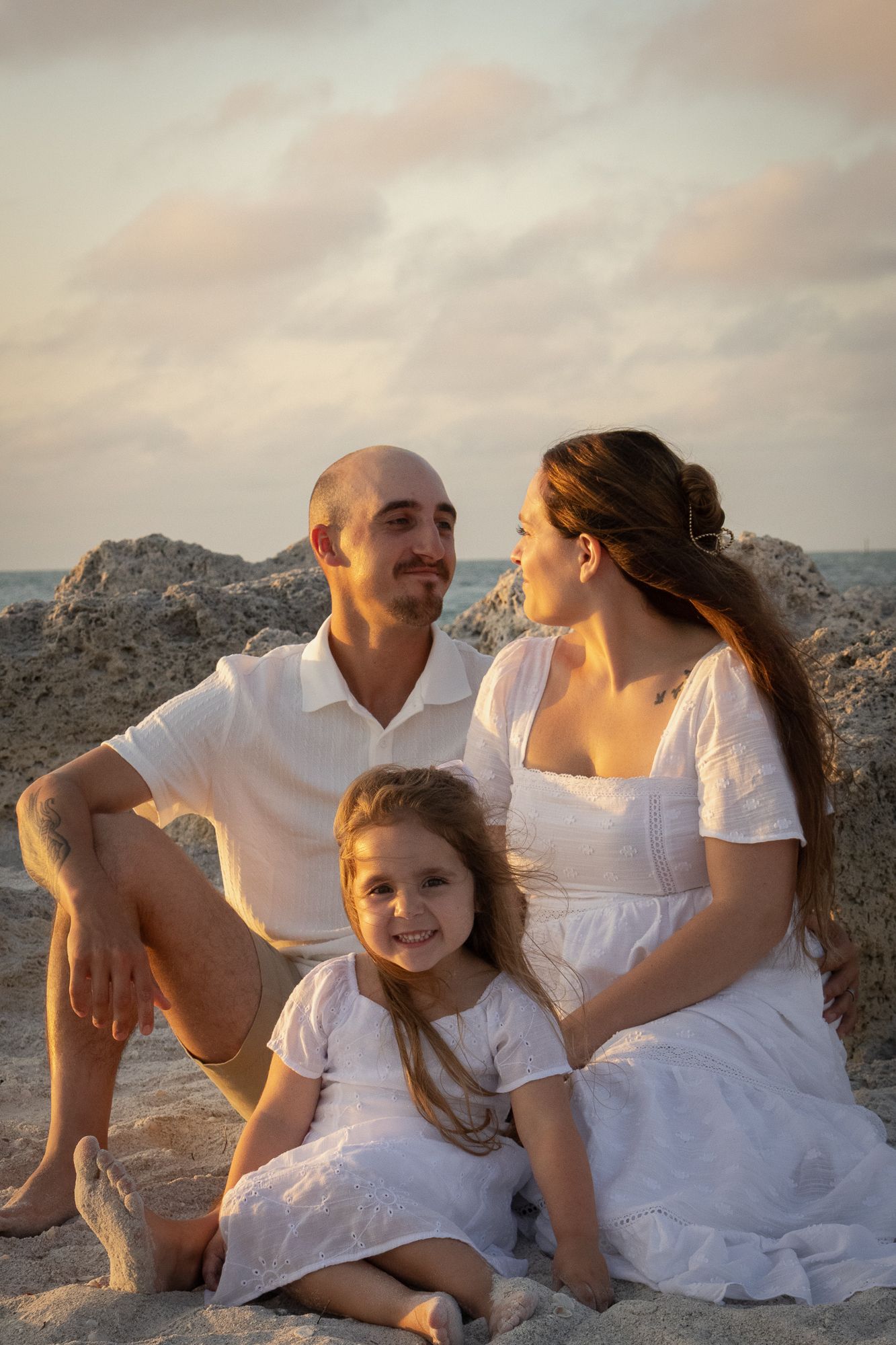 Family of three in white outfits sitting on beach rocks at golden sunset.