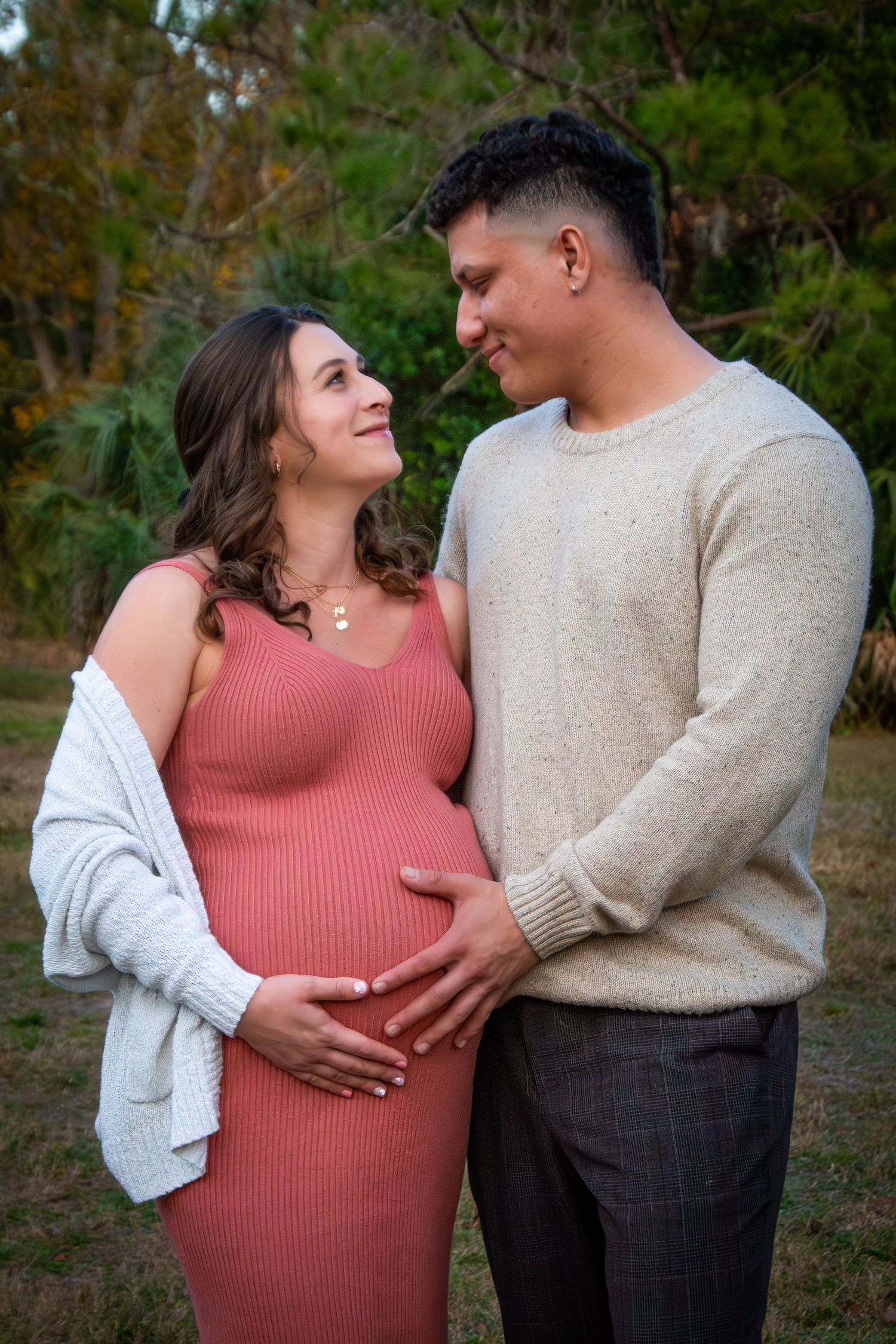 Pregnant couple in a park, woman in pink dress holding baby bump, man in beige sweater.