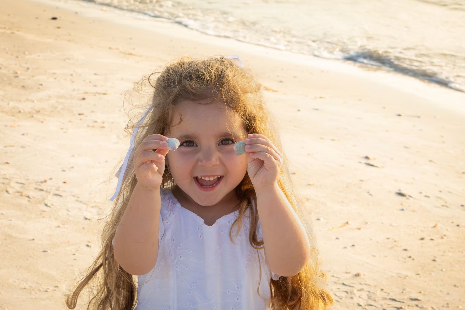 Joyful little girl in white dress holding shells on sunny sandy beach.