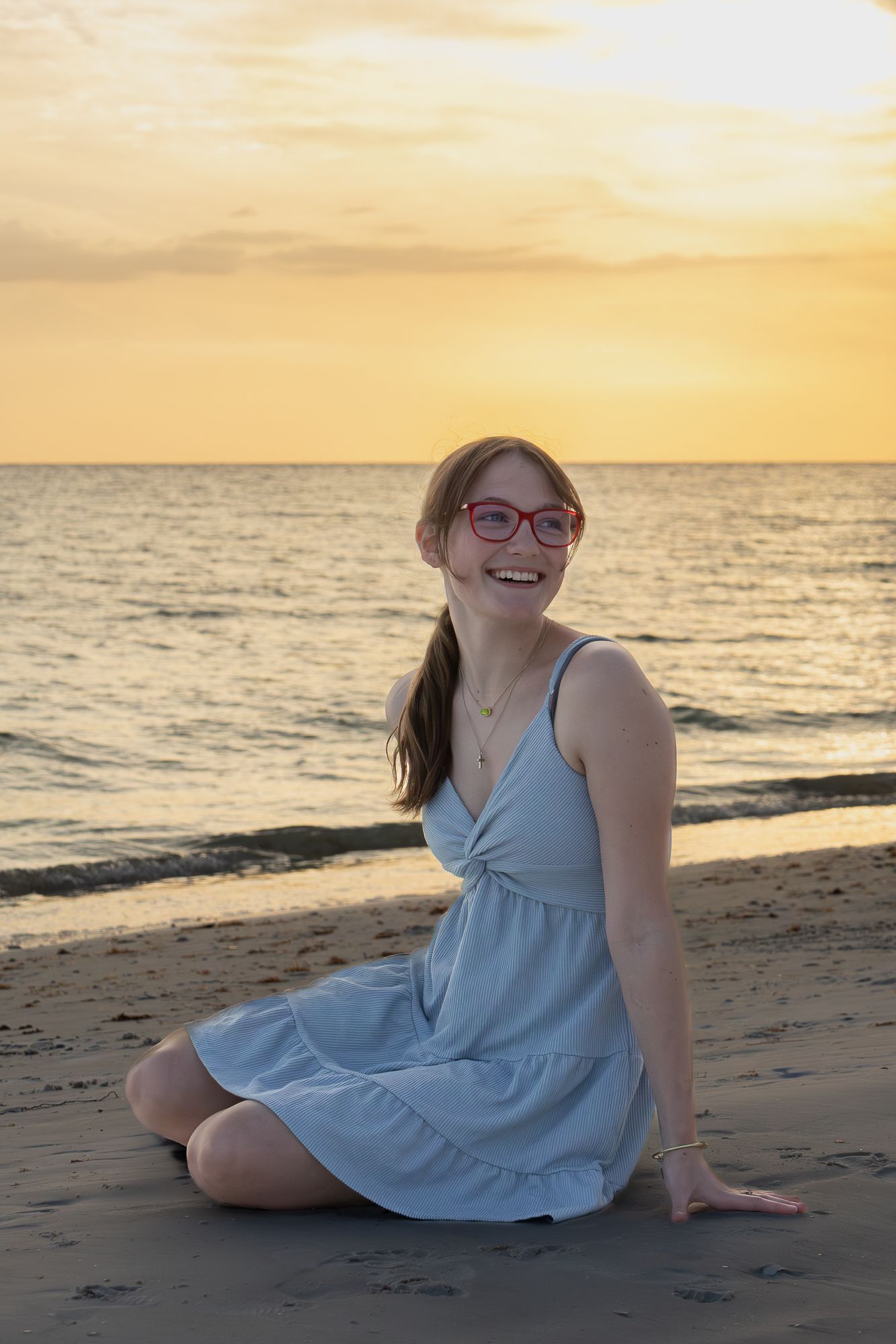 Smiling teenage girl with red glasses sitting on sandy beach in blue dress at golden sunset.