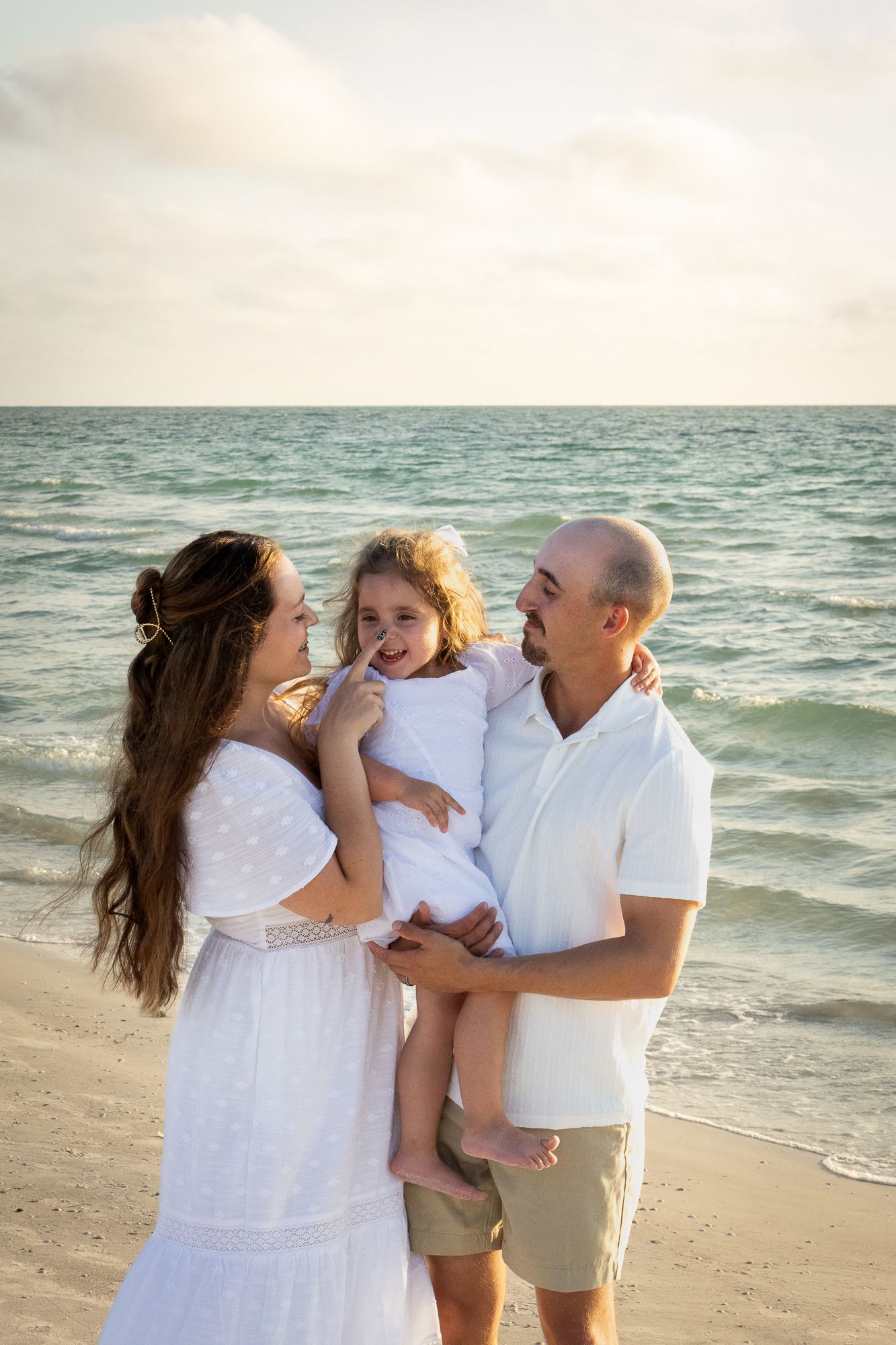Happy family of three dressed in white sharing a tender moment together on a beautiful beach at sunset.