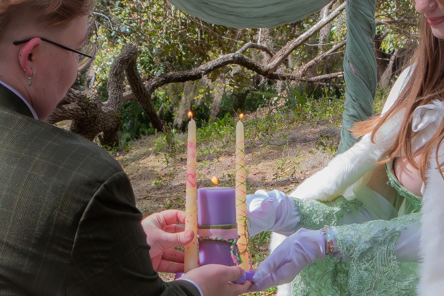 Couple lighting unity candle together during outdoor wedding ceremony under tree arch.