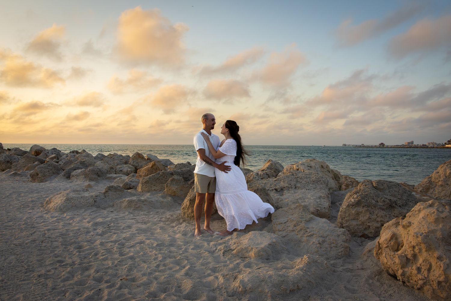 A romantic couple embraces on a rocky beach at sunset, the woman in a flowing white dress with golden clouds above.