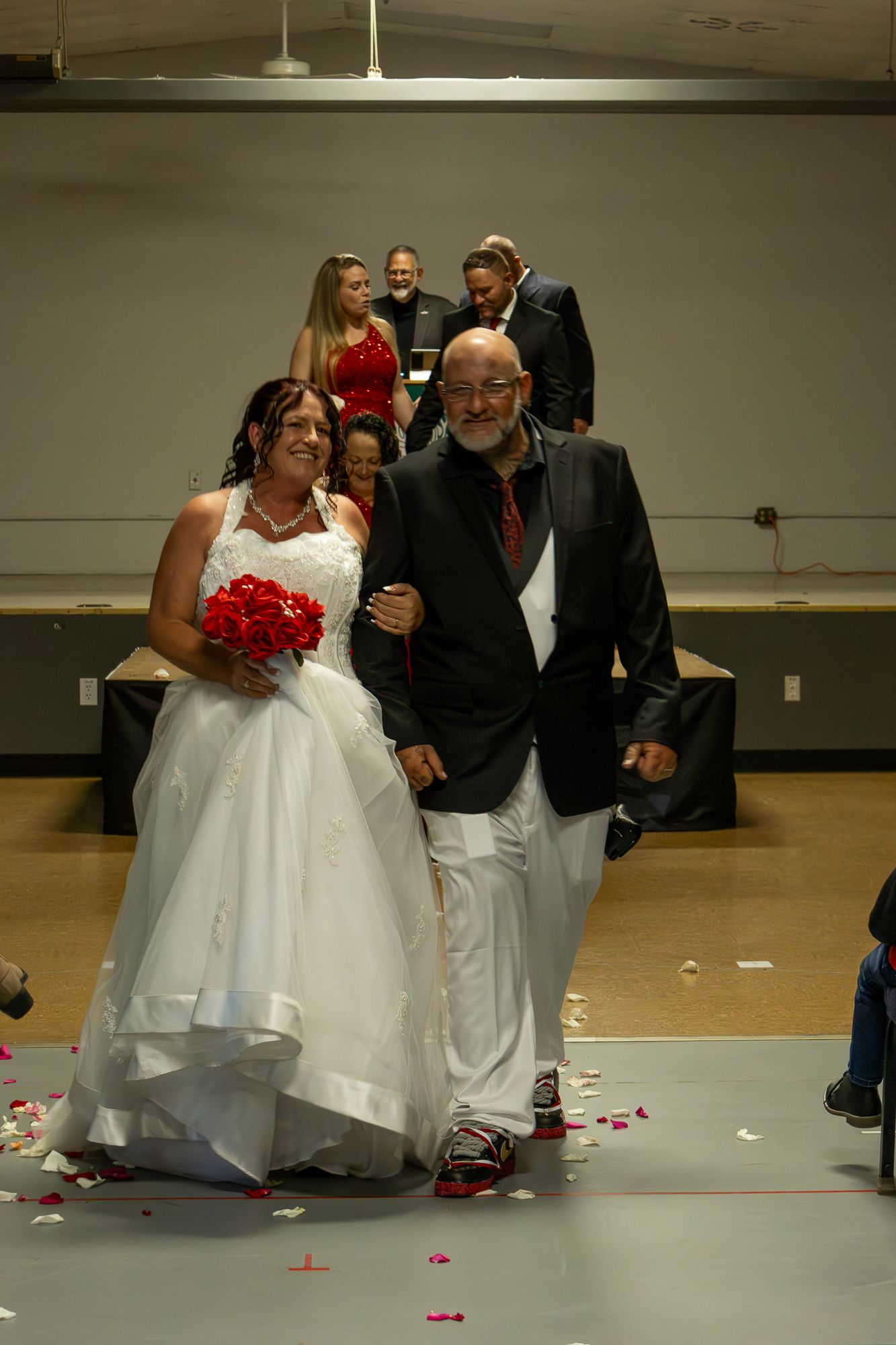 Bride in white gown holding red bouquet walks down aisle with escort at indoor wedding ceremony.