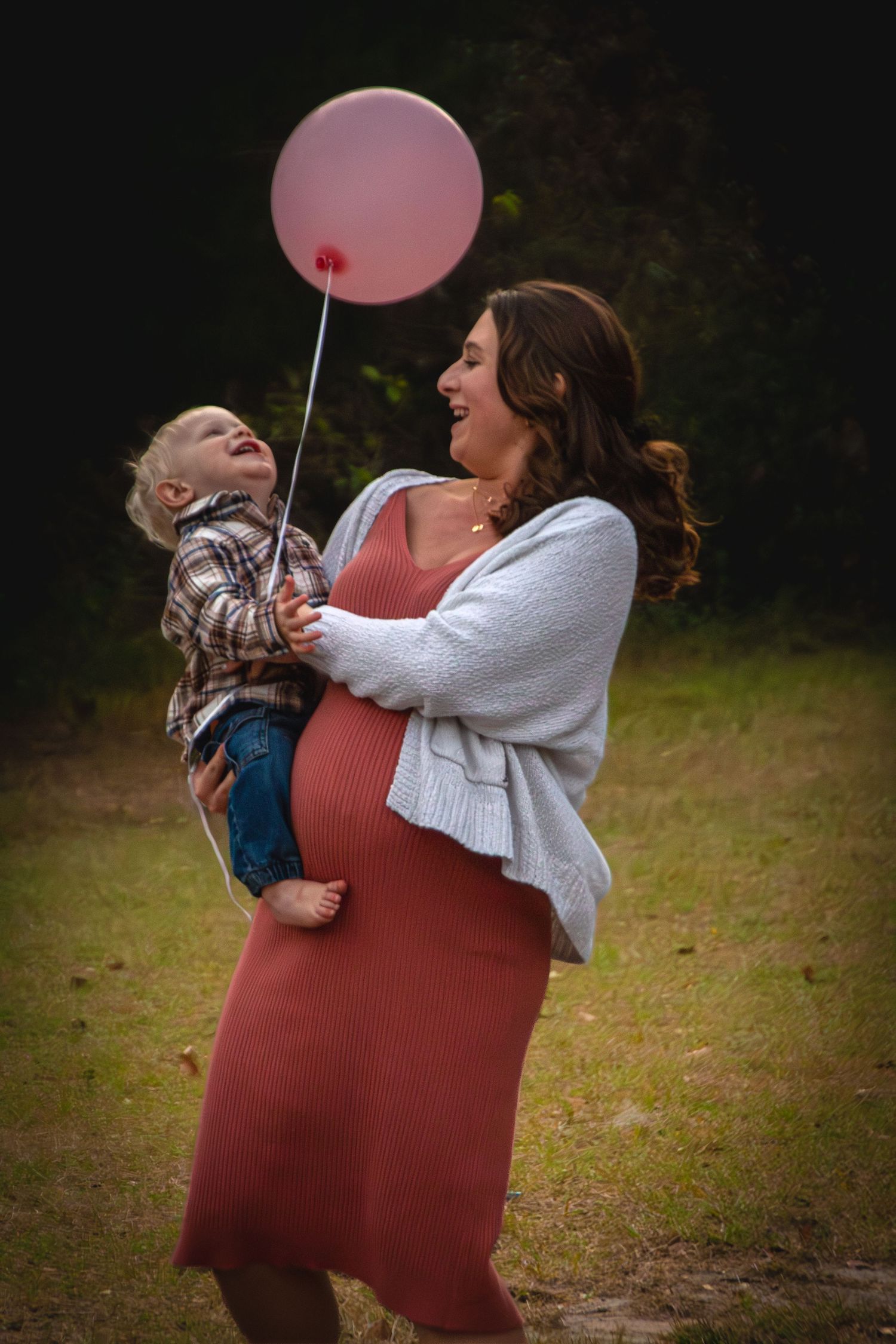 Pregnant woman in pink dress holding toddler and pink balloon in a park.