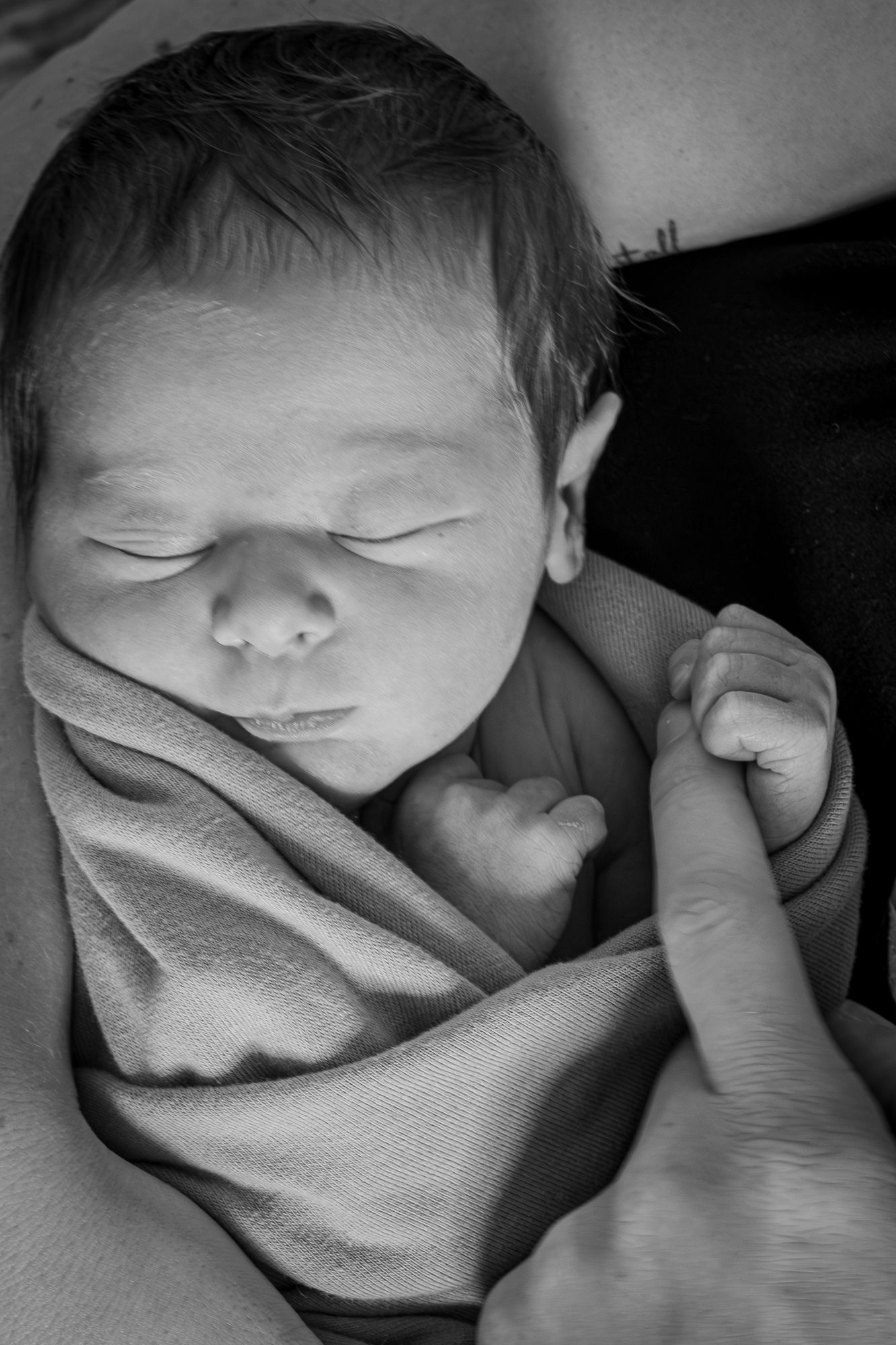Newborn baby sleeping peacefully wrapped in a blanket, grasping an adult finger in black and white photo.