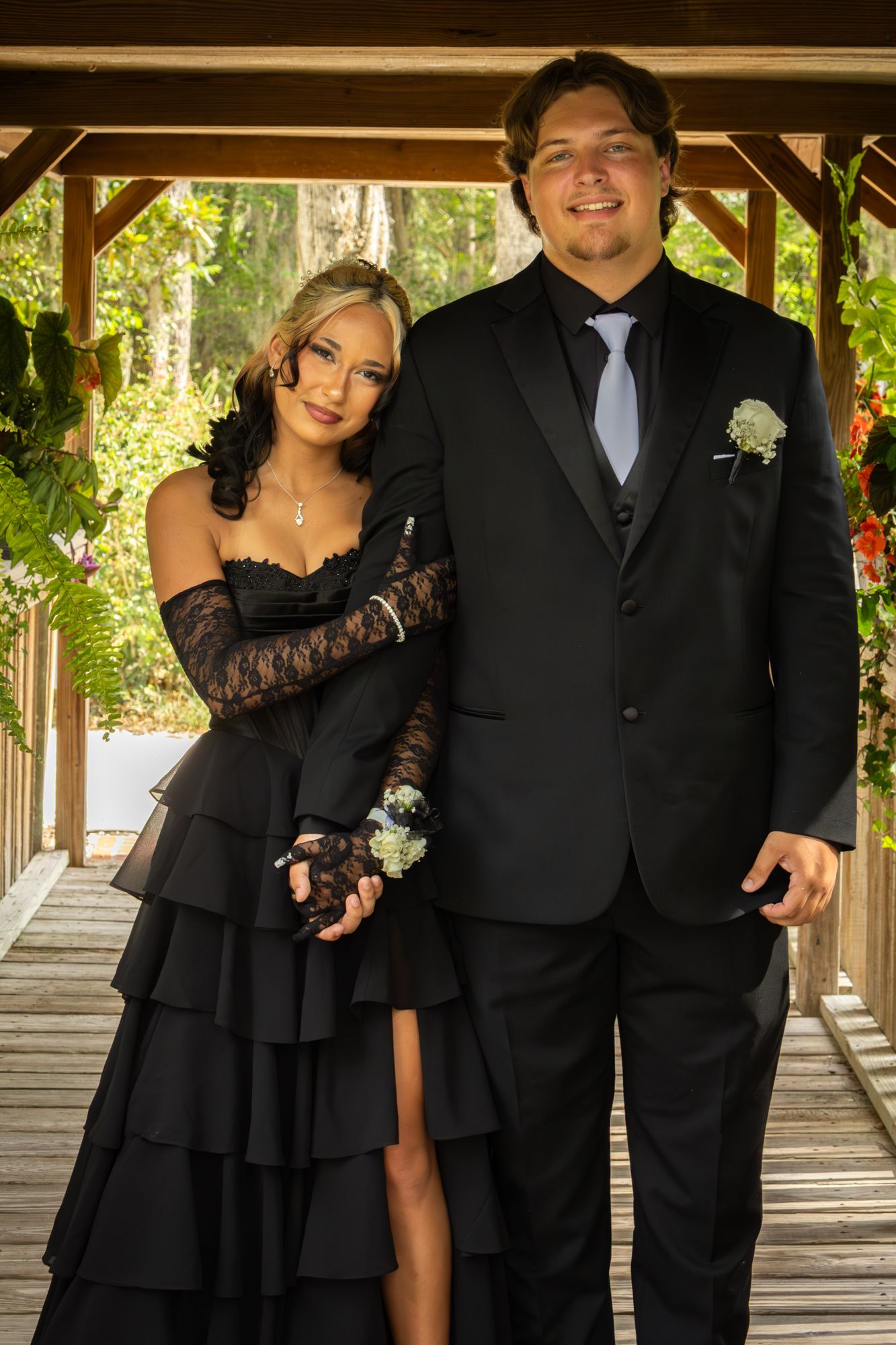 A stylish couple stands under a wooden gazebo, the woman in a layered black dress with lace gloves and a corsage.