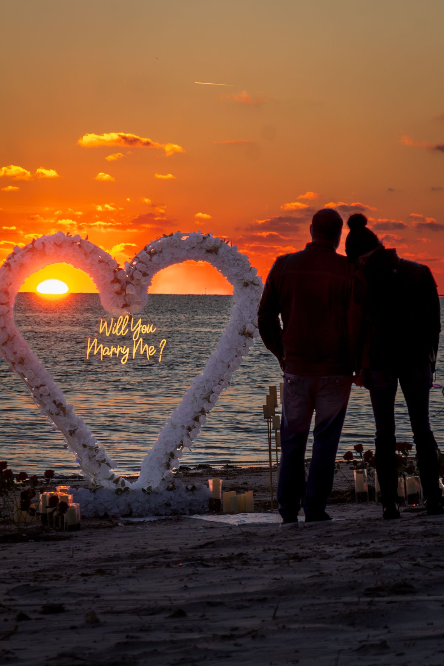 A couple silhouetted against a vibrant sunset beside a floral heart arch reading Will You Marry Me.