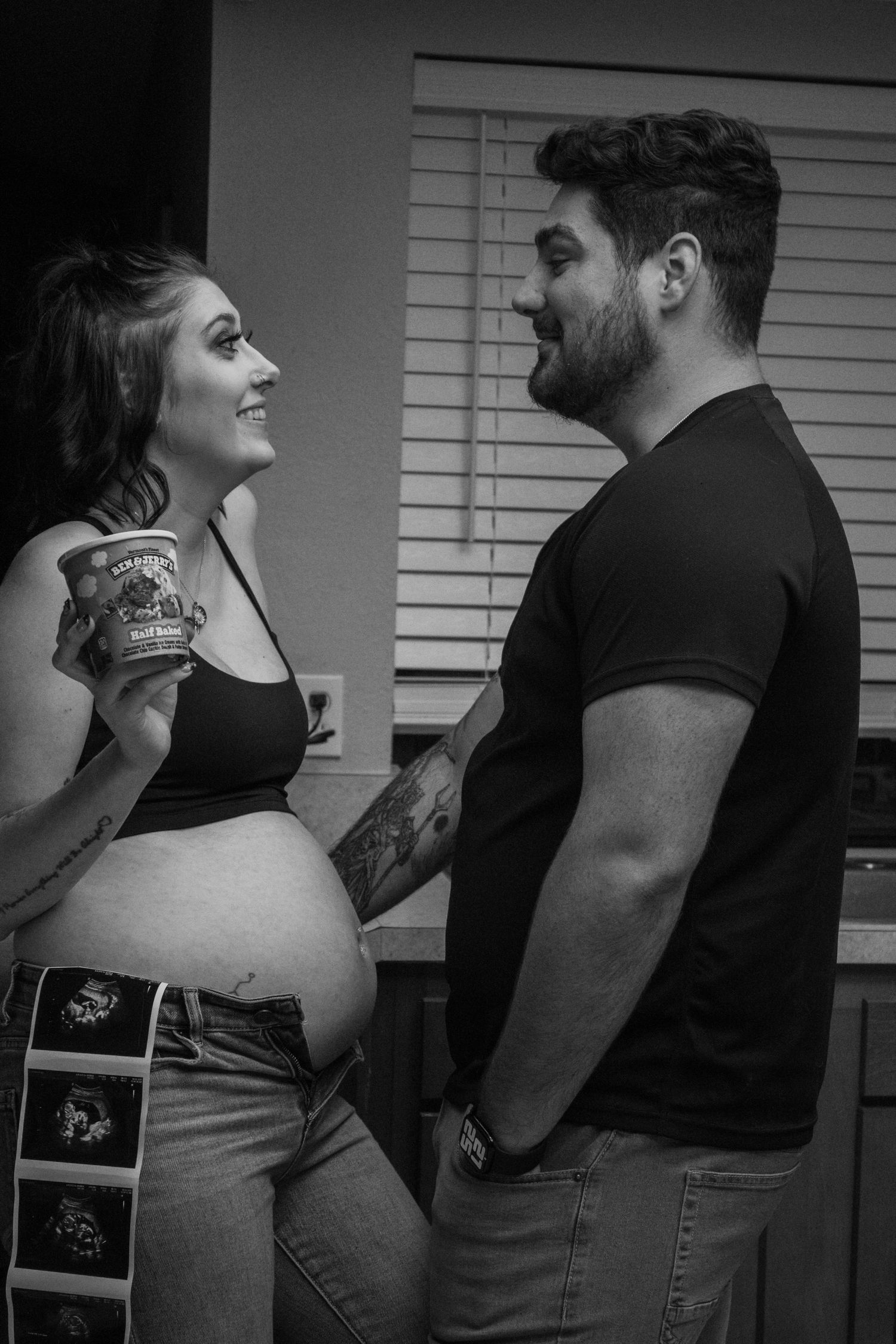 Black and white photo of a smiling pregnant woman holding ice cream with a tattooed man, ultrasound photos tucked in her waistband.