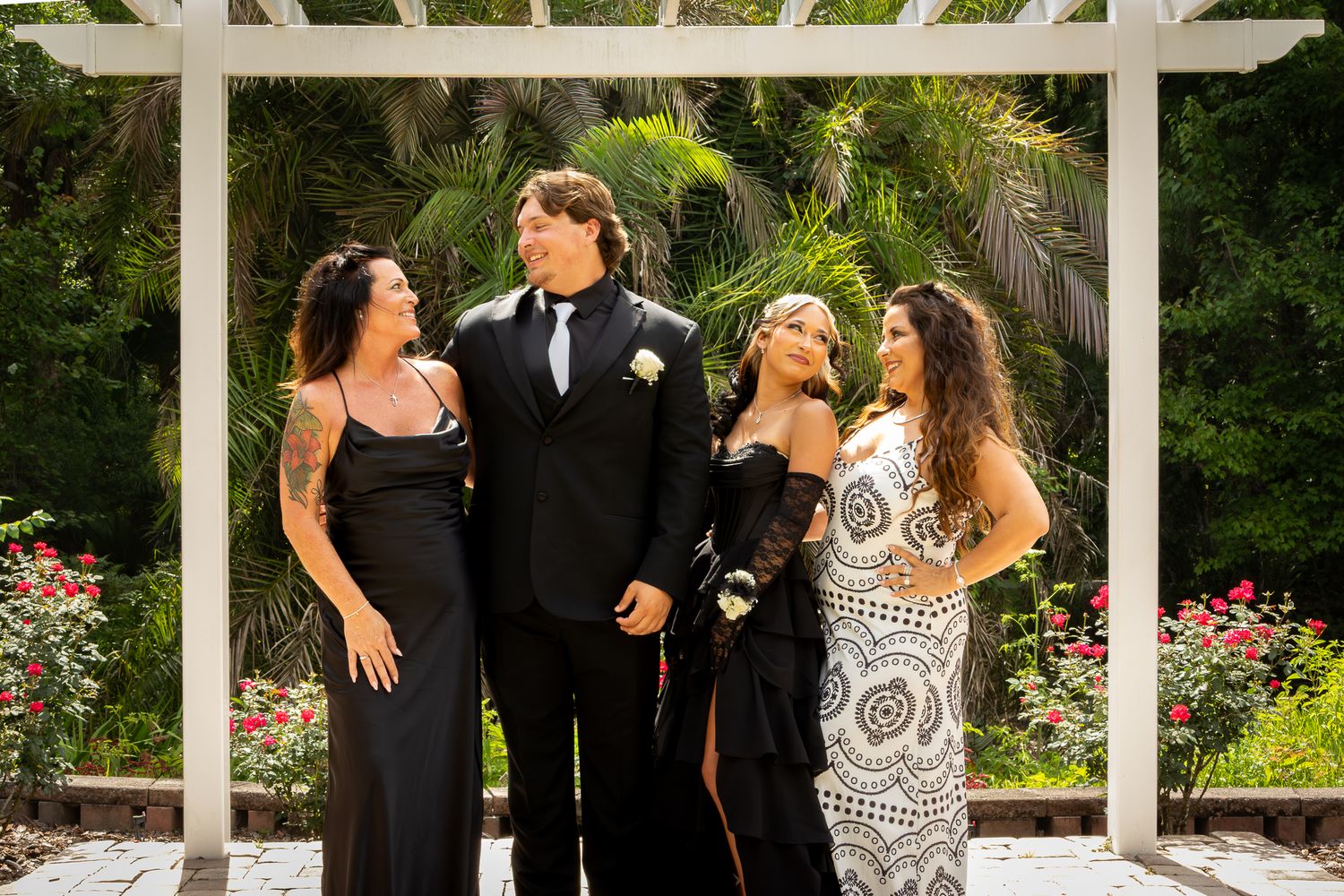 Group of four people dressed formally in black and white prom attire posing under white pergola.