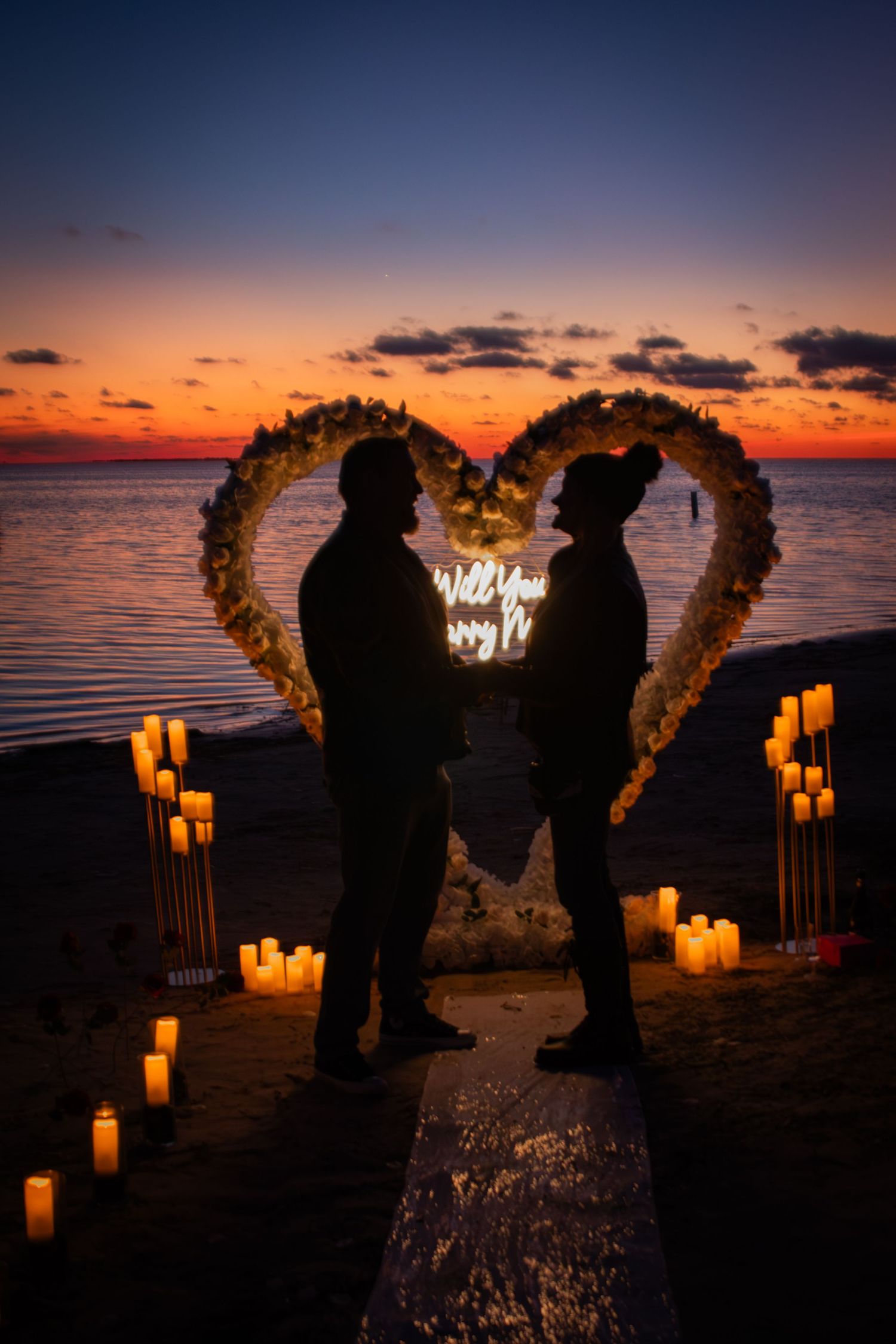 A couple stands silhouetted inside a floral heart arch surrounded by candles at a beach sunset.