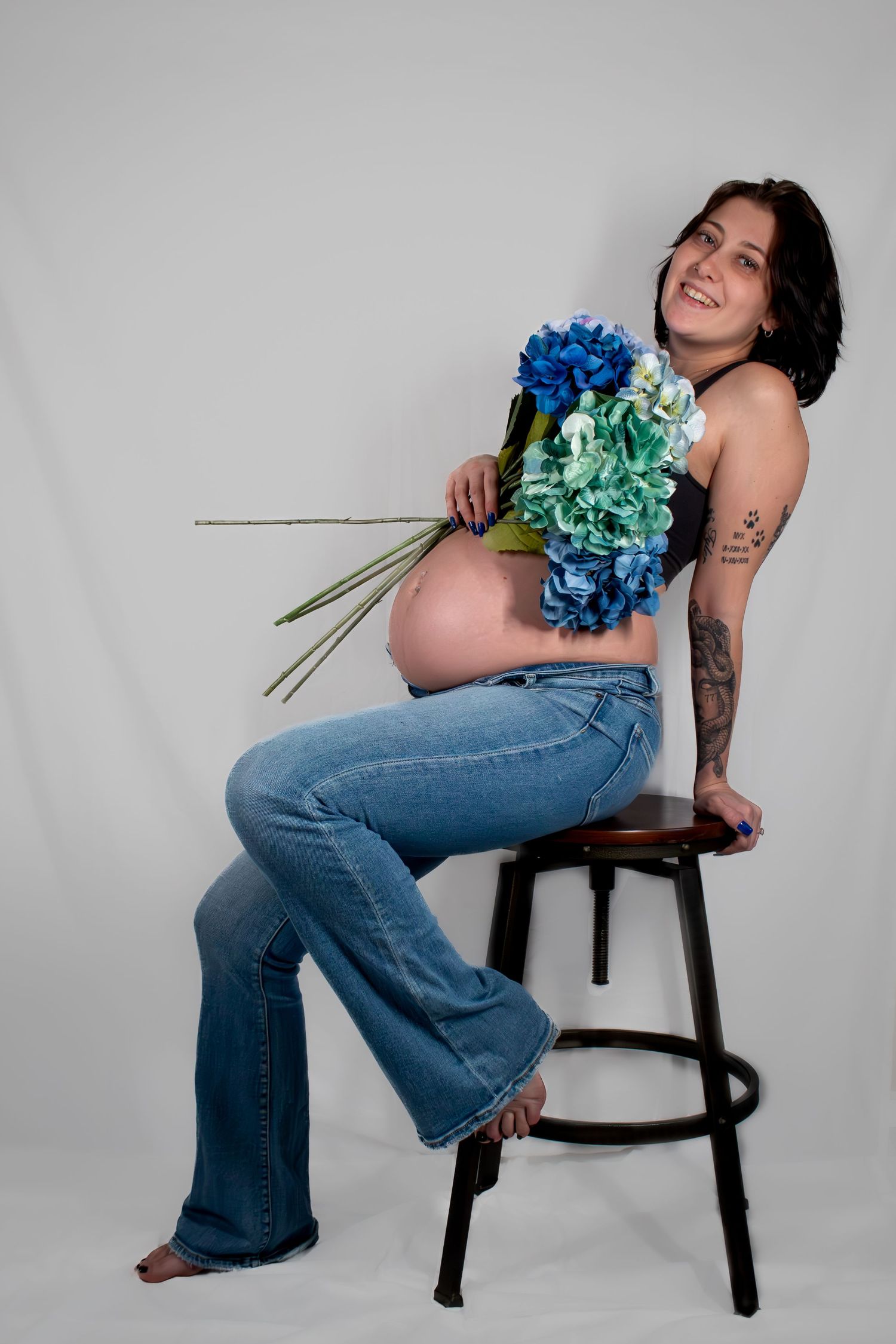 A smiling pregnant woman sits on a stool holding blue hydrangeas, wearing jeans in a studio.