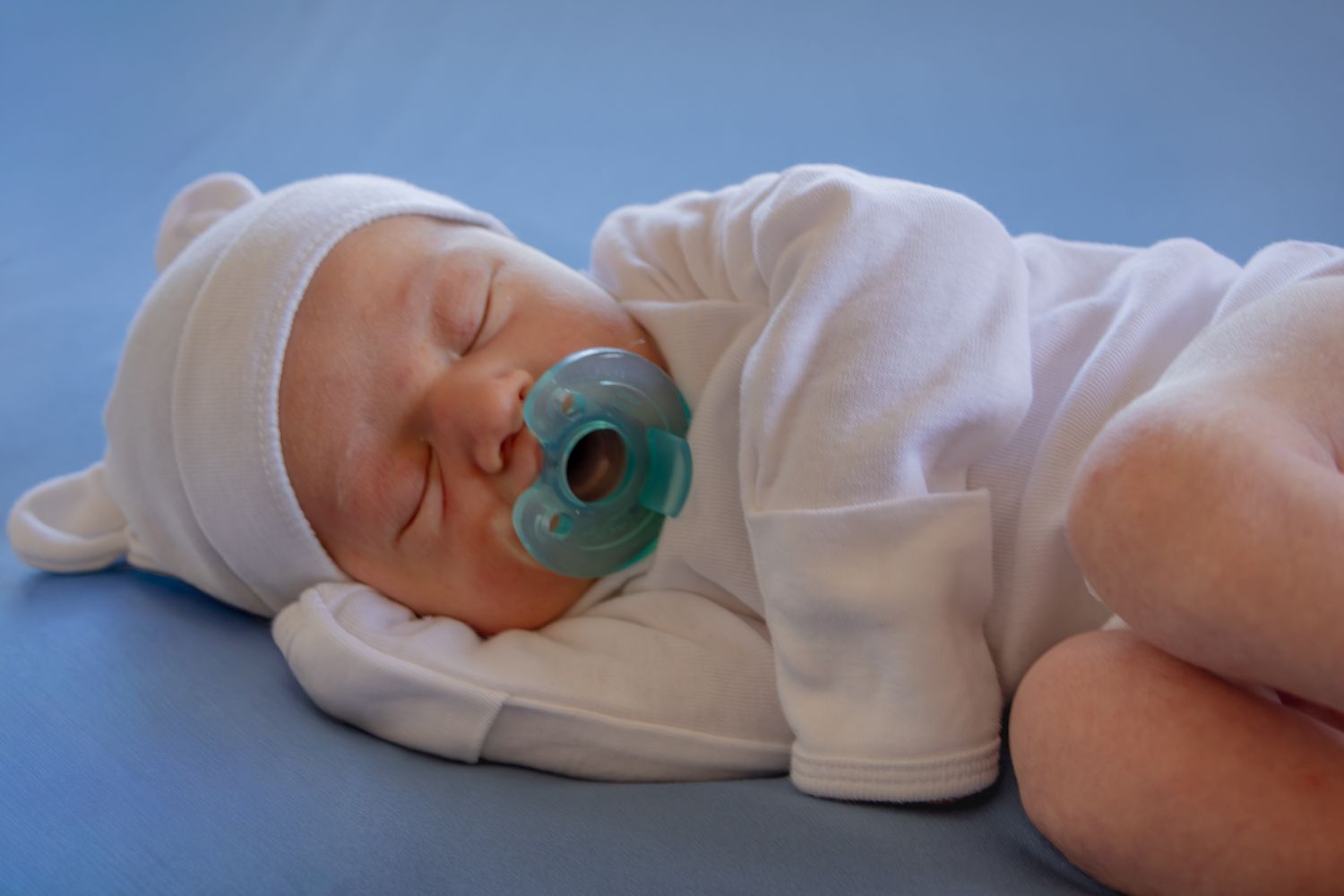 Peaceful newborn baby sleeping in white outfit and hat with a blue pacifier on a blue background.