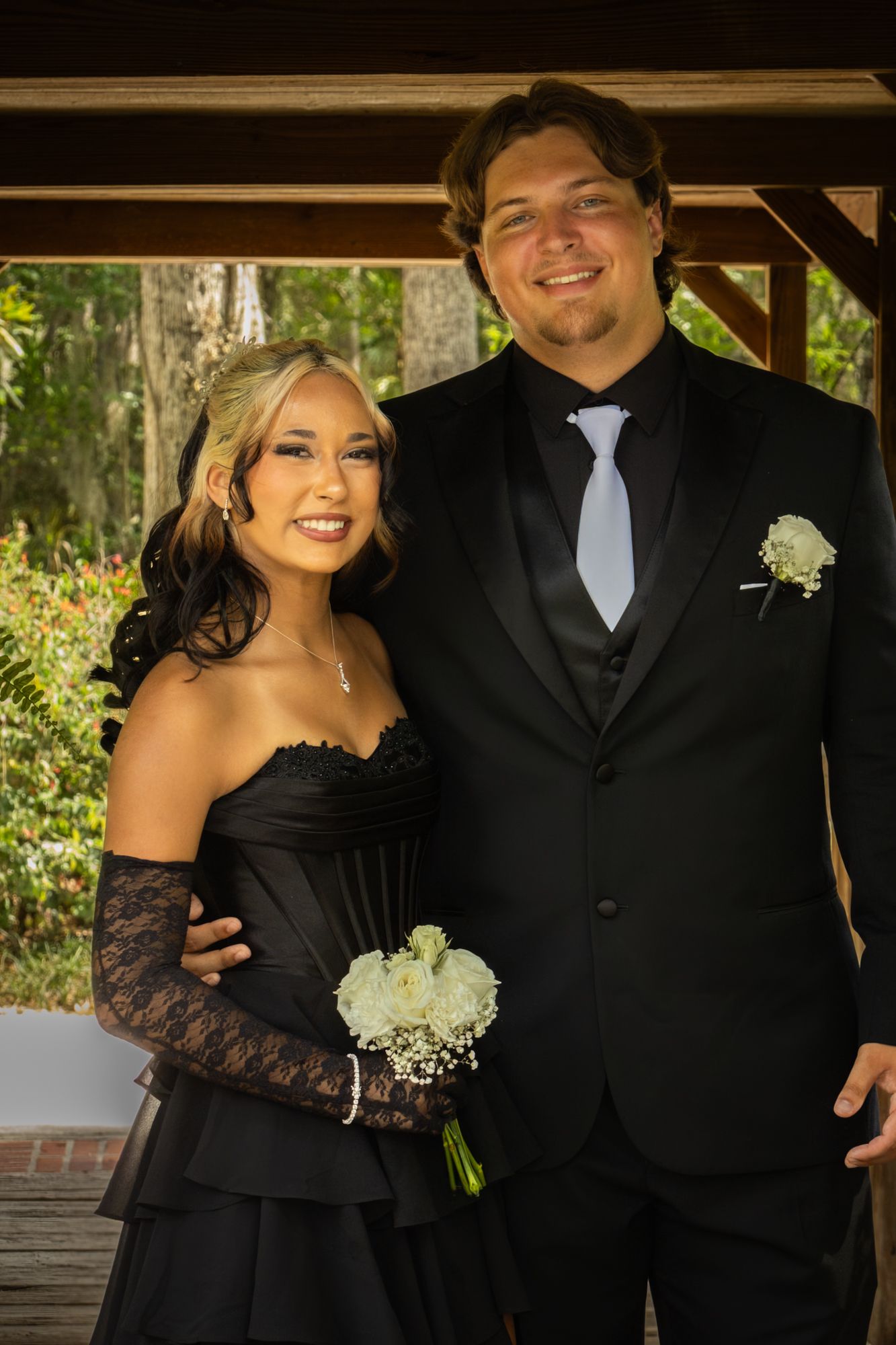 An elegant couple poses under a gazebo, the woman in a black strapless gown holding white roses, the man in a suit.