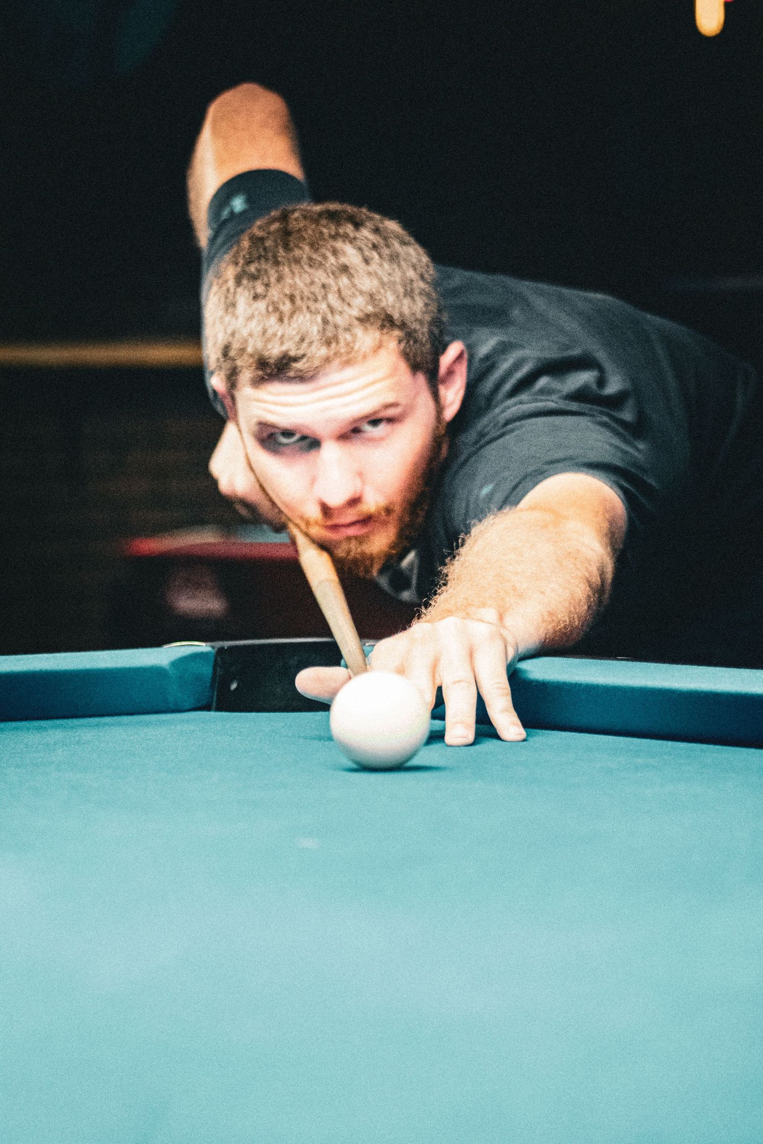 A focused young man with a beard leans over a pool table, aiming his cue stick at the white cue ball.