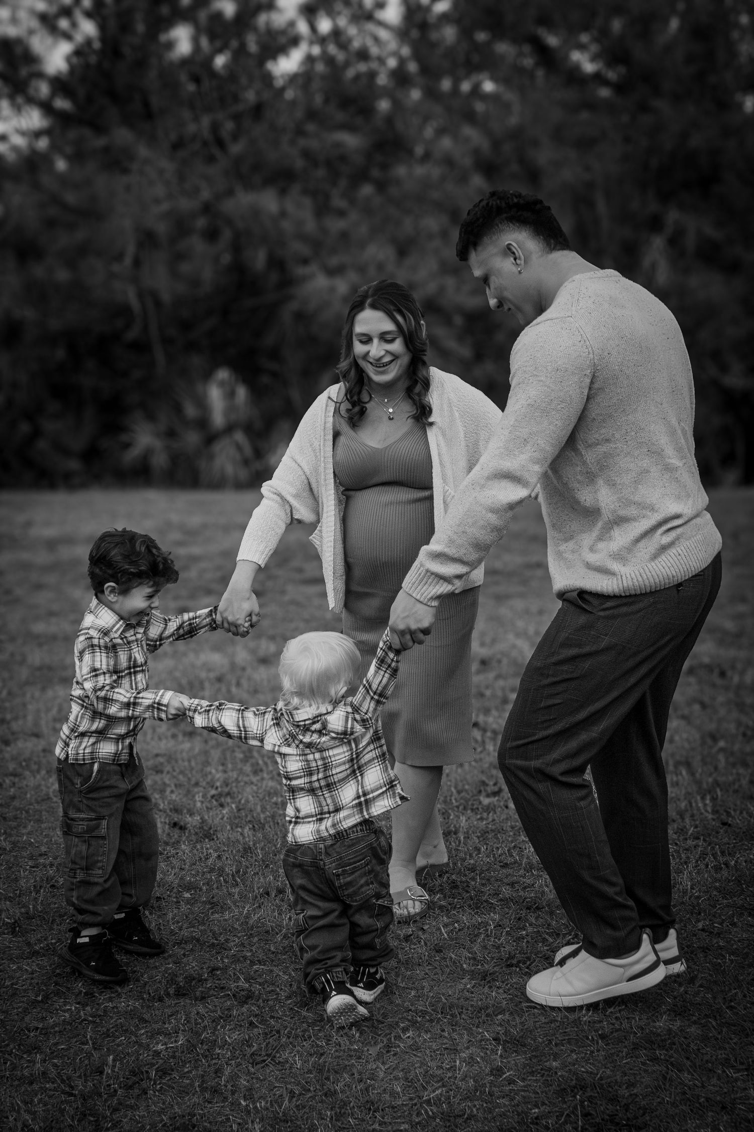 Black and white photo of pregnant family holding hands with two young children outdoors.