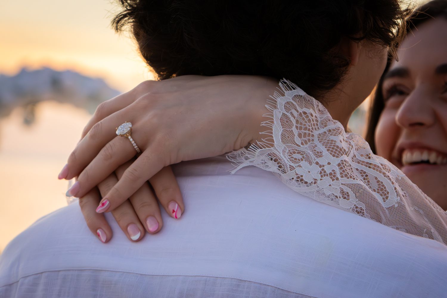 Bride showing diamond engagement ring while embracing groom at sunset beach.
