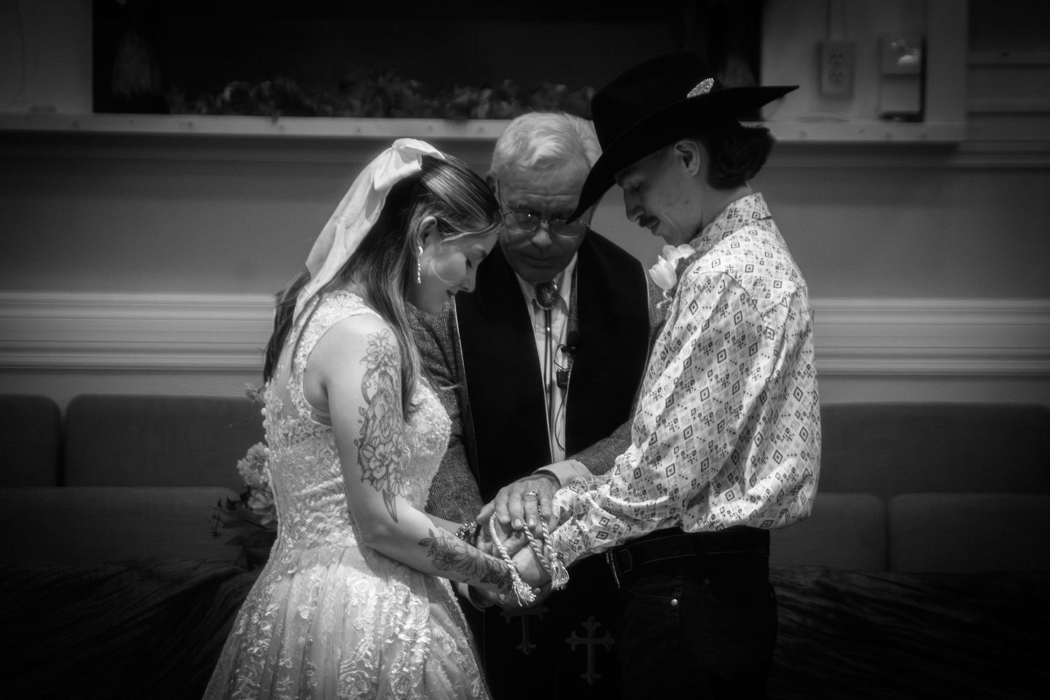 Black and white photo of bride and groom holding hands in prayer with officiant at altar.