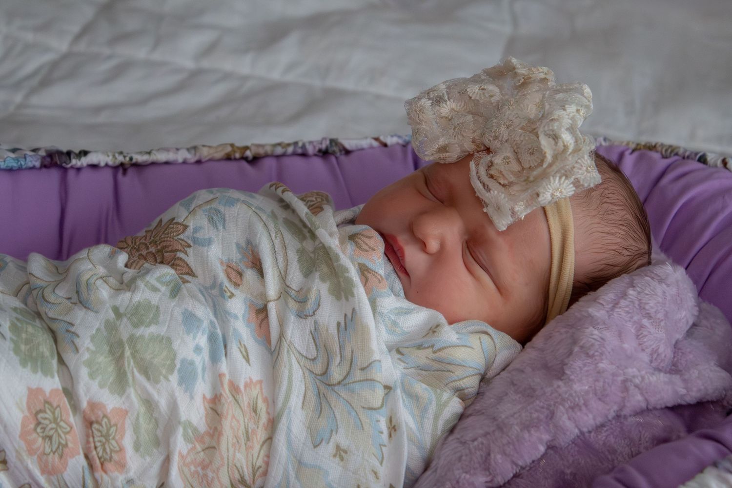 Sleeping newborn baby swaddled in floral blanket with lace headband on purple bedding.