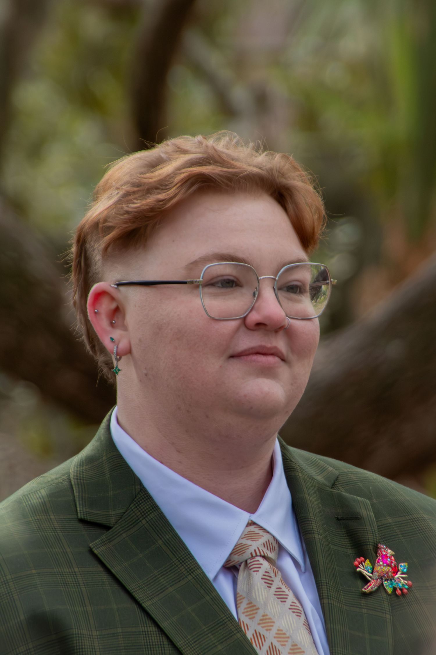 Person with red hair and glasses wearing a green blazer with a floral brooch outdoors.