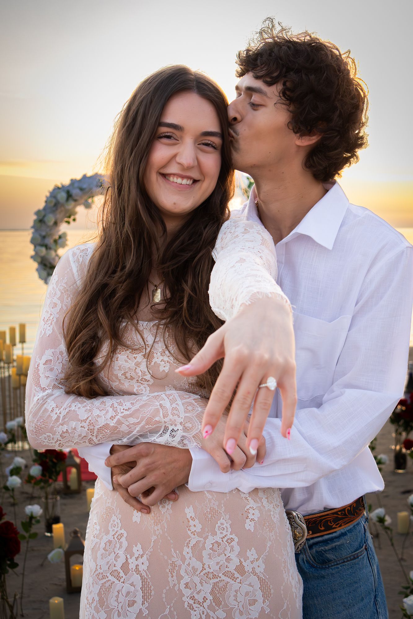 Smiling couple displaying engagement ring at beach sunset proposal with floral arch.