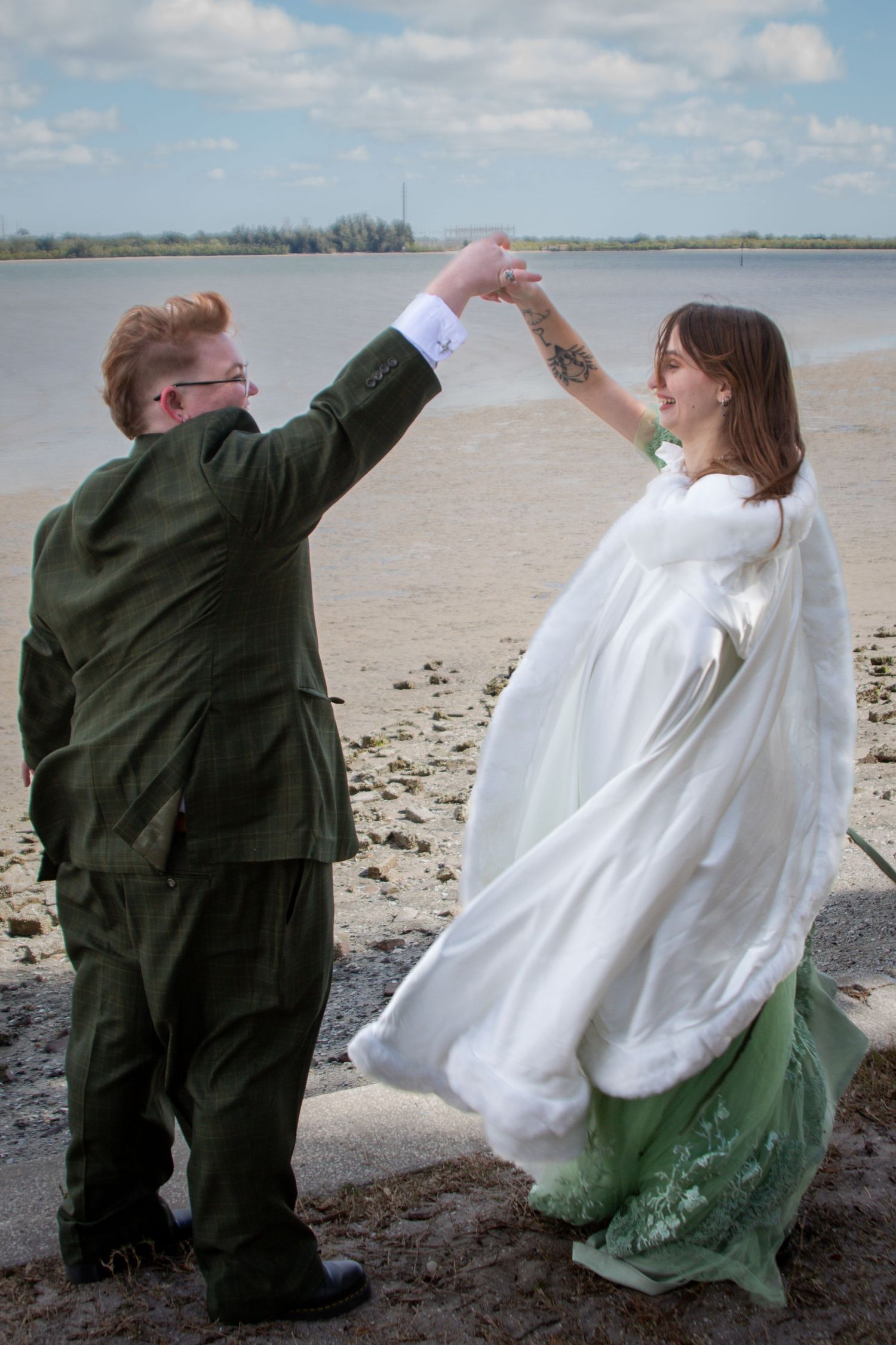 Newlyweds dancing on sandy beach, bride in white cape and green dress, groom in olive suit.