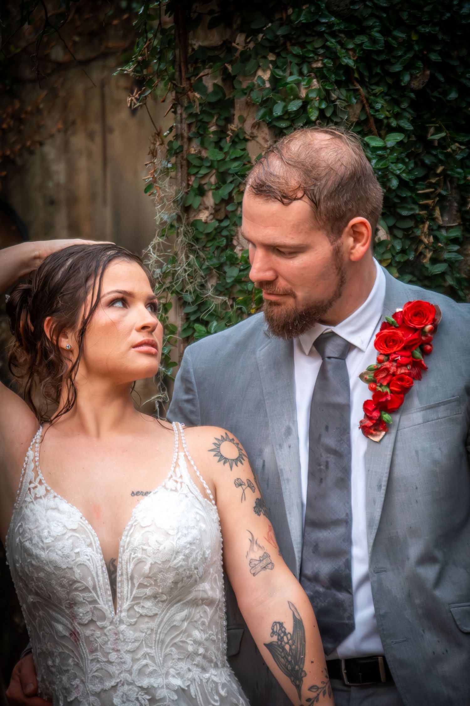 Tattooed bride and bearded groom in gray suit share intimate gaze against ivy-covered stone wall.