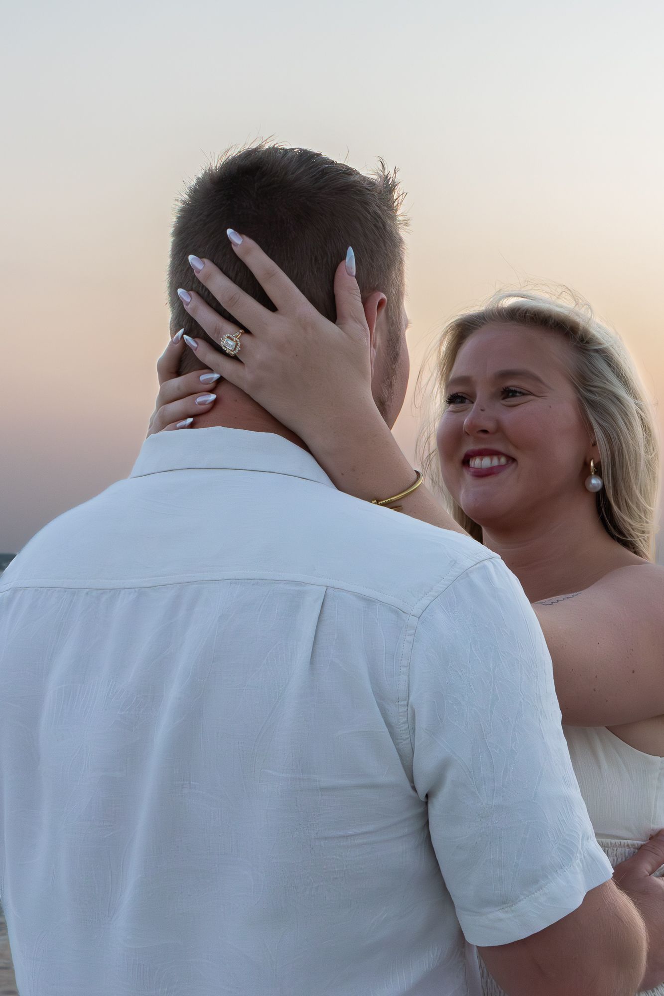 Newly engaged woman showing off her diamond ring while embracing her partner at sunset on the beach.