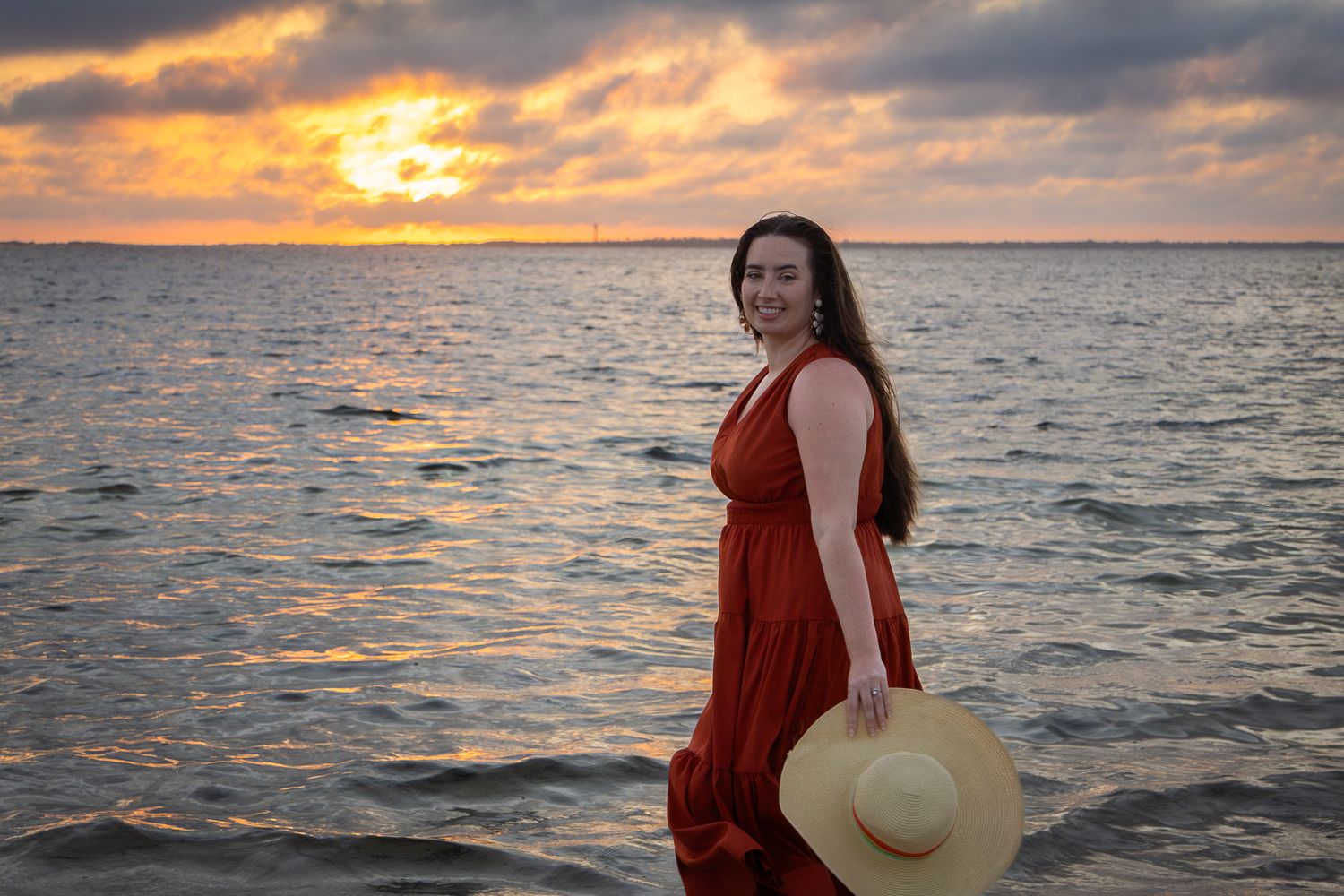 Woman in red dress holding a straw hat posing by the ocean during a vibrant golden sunset.
