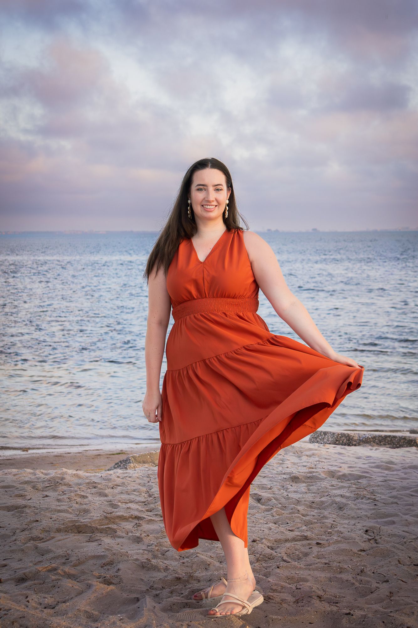 Woman in flowing red dress posing on sandy beach at colorful sunset.