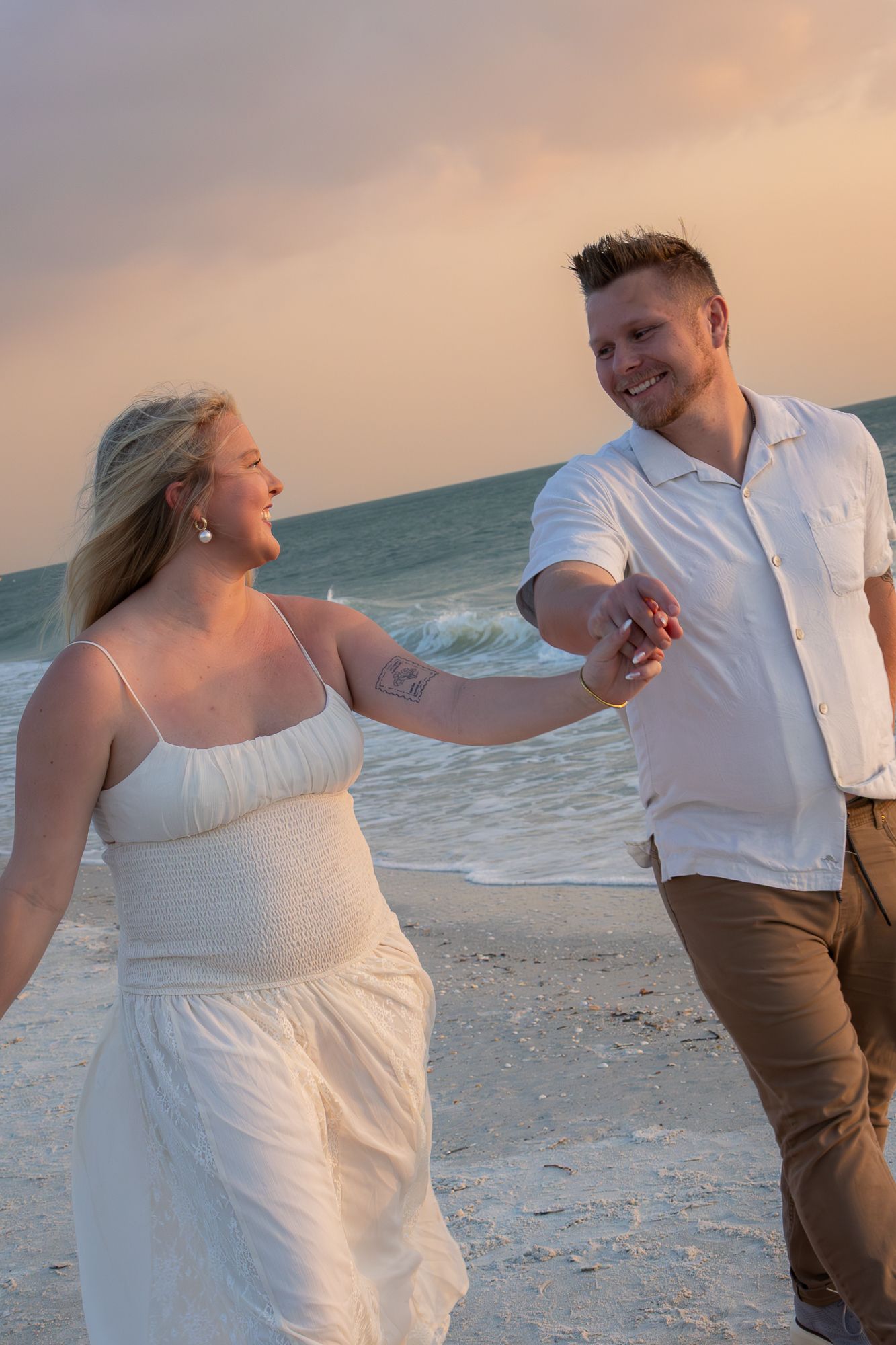 Joyful couple holding hands and laughing together while walking on a sandy beach at golden sunset.