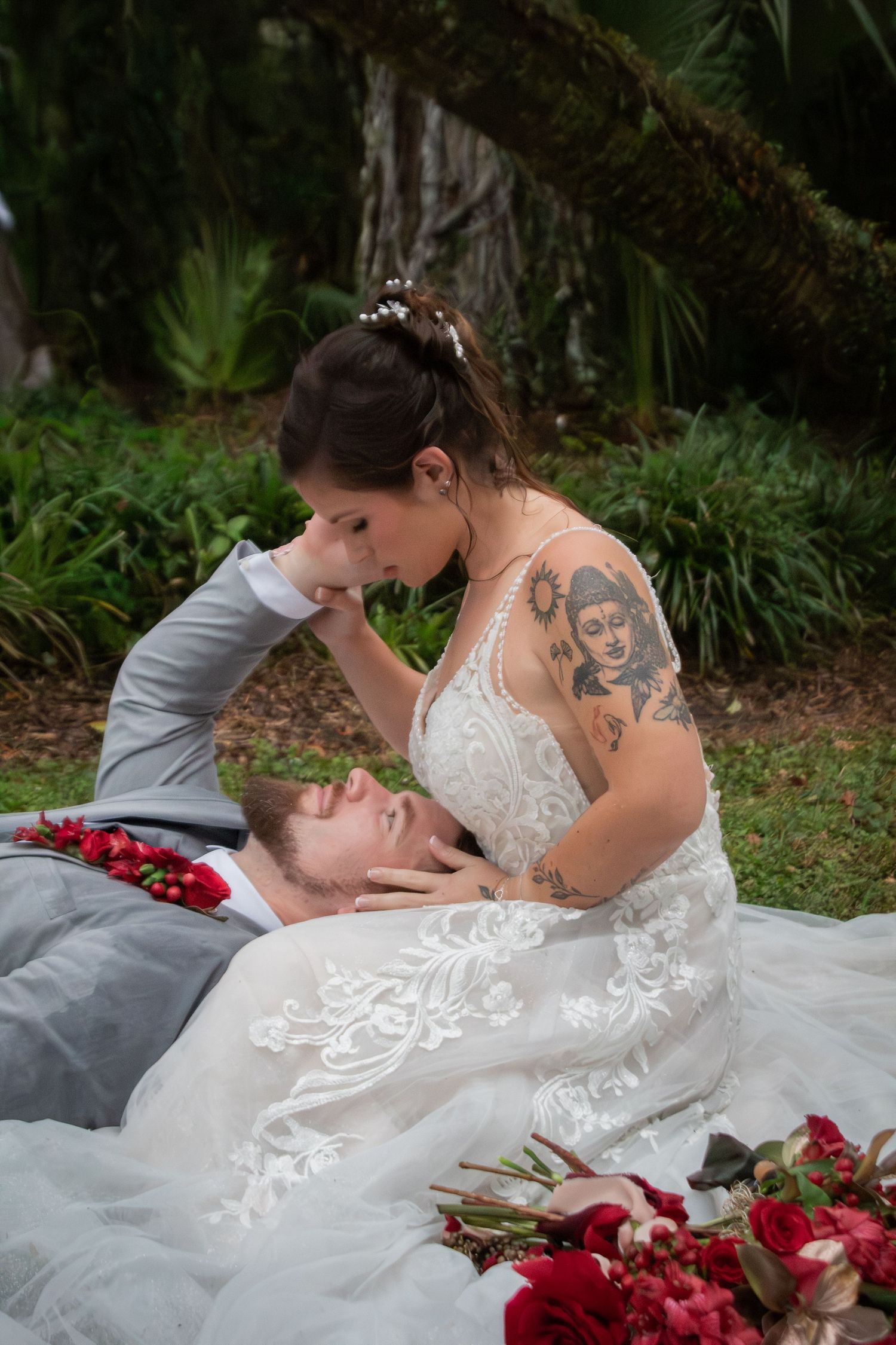 Bride in lace wedding gown tenderly holds grooms face while he lies among red rose petals.