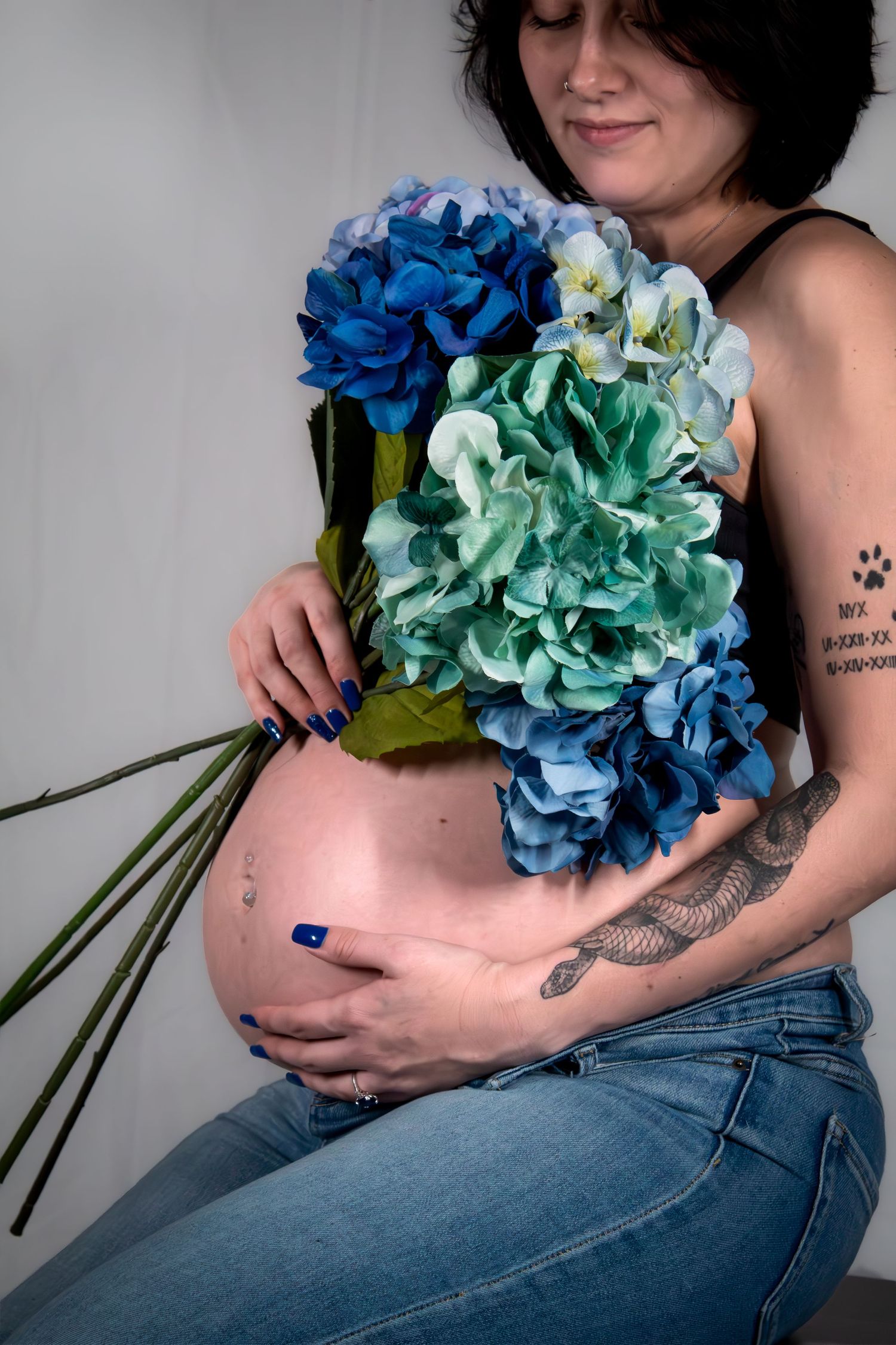 Pregnant woman holding blue hydrangea bouquet against bare belly, wearing jeans, showing tattoo on arm.