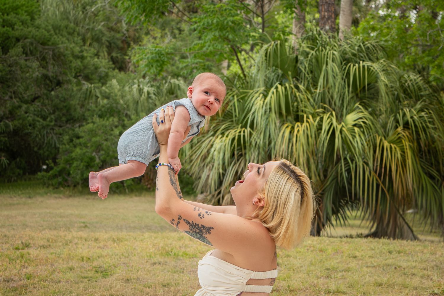 Blonde mother joyfully lifting her laughing baby in the air outdoors with green trees in background.