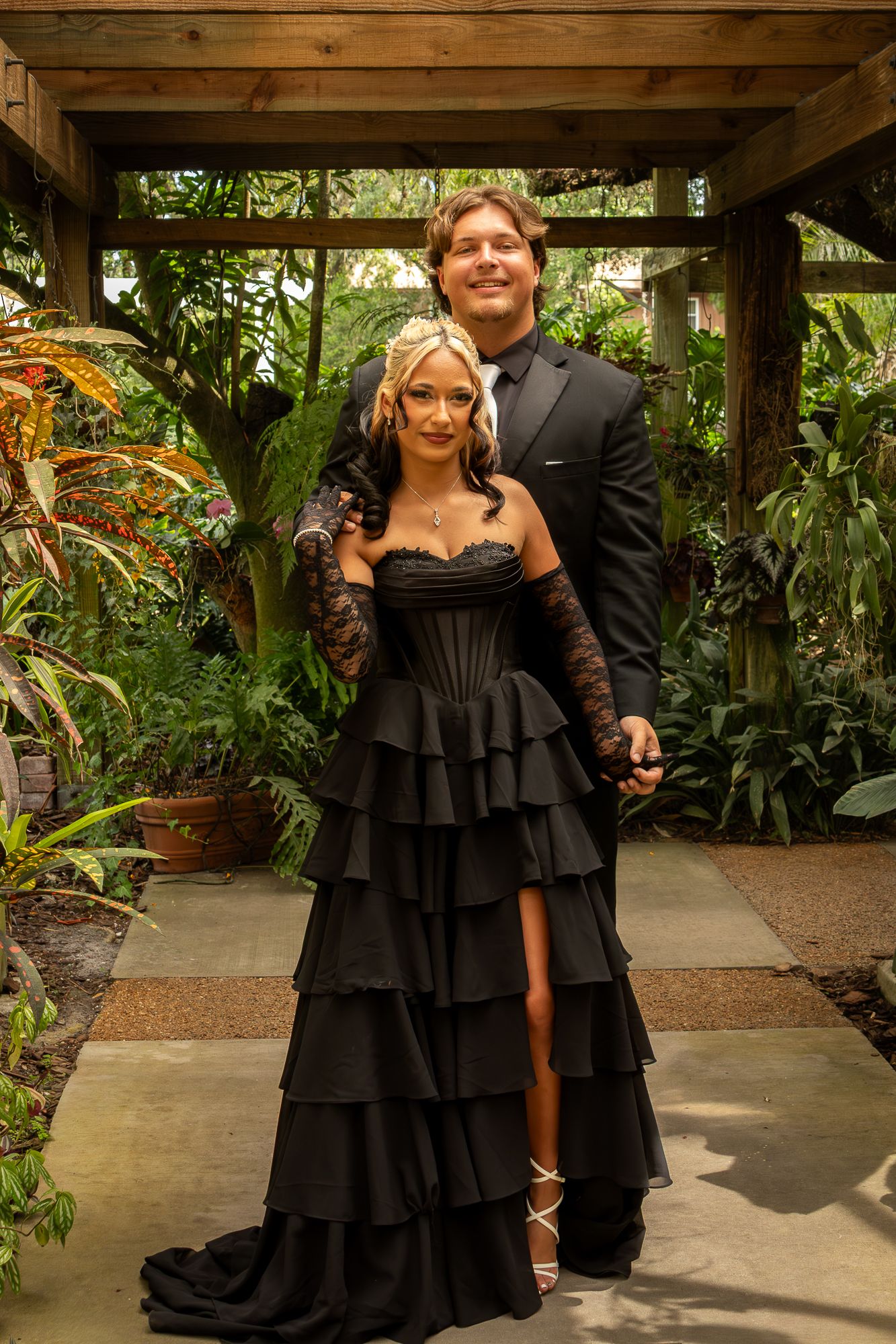 A couple poses in a lush garden gazebo, the woman in a tiered black gown with lace gloves, the man in a black suit.