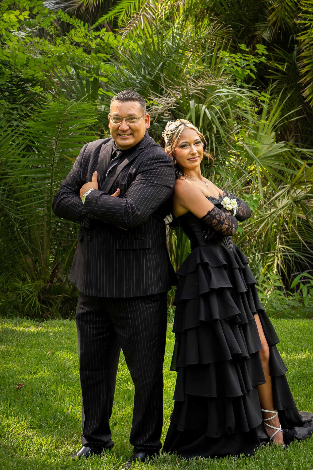 Man in pinstripe suit and woman in black ruffled prom dress pose together in lush garden setting.