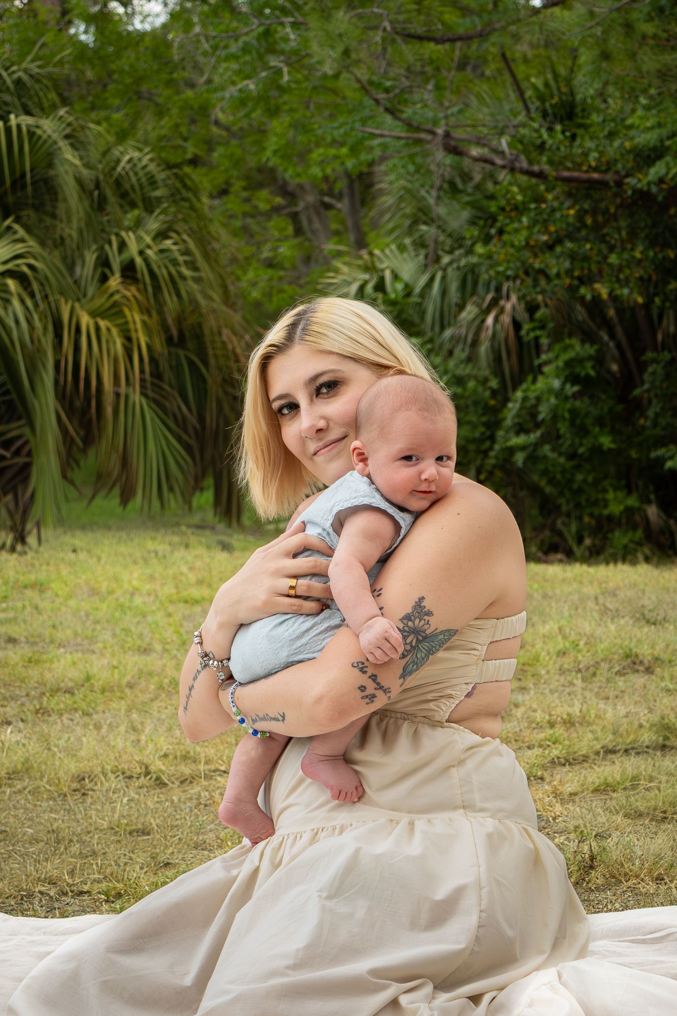 Young blonde mother with tattoos in cream dress holds newborn baby outdoors in lush green park setting.