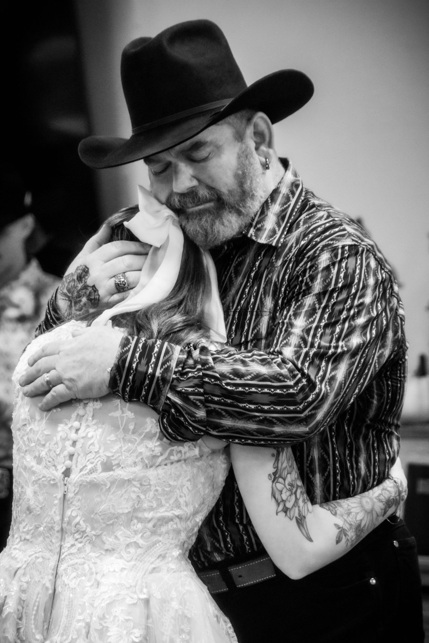 Black and white photo of the father in a cowboy hat sharing an emotional embrace with his daughter in a wedding dress.